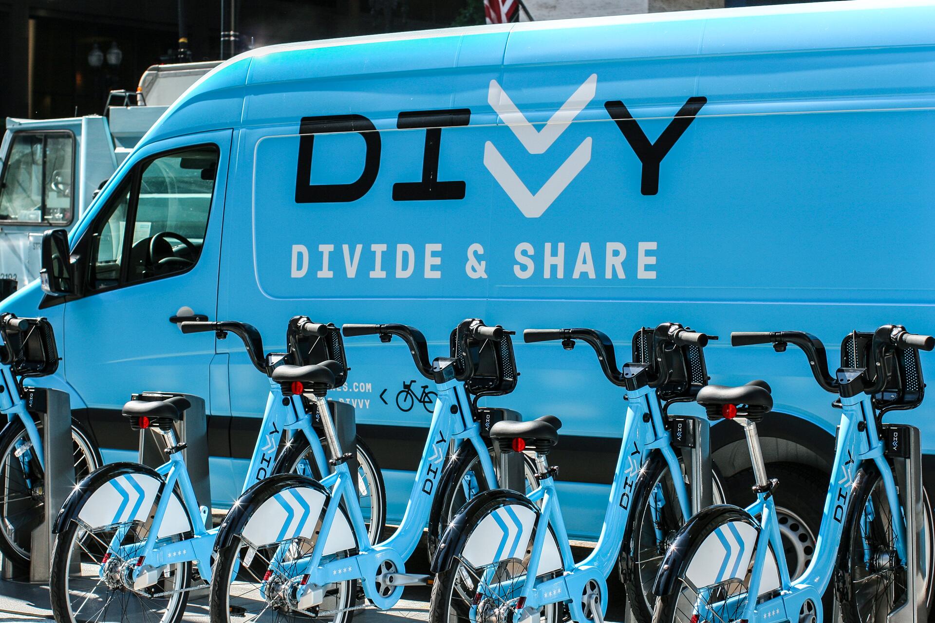 Blue van labeled "DIVVY Divide & Share" with several blue bicycles parked in front, featuring a matching design with white arrow patterns.