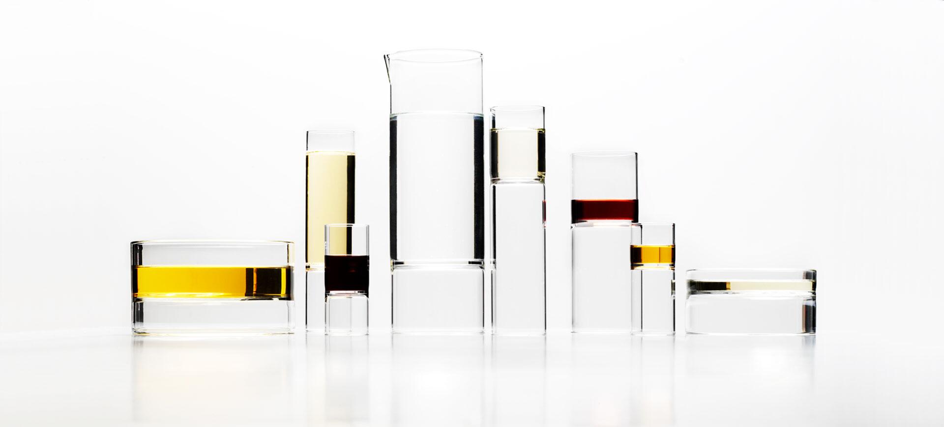 Glass containers of varying heights with colored liquids, including yellow, red, and clear substances, arranged against a white background.