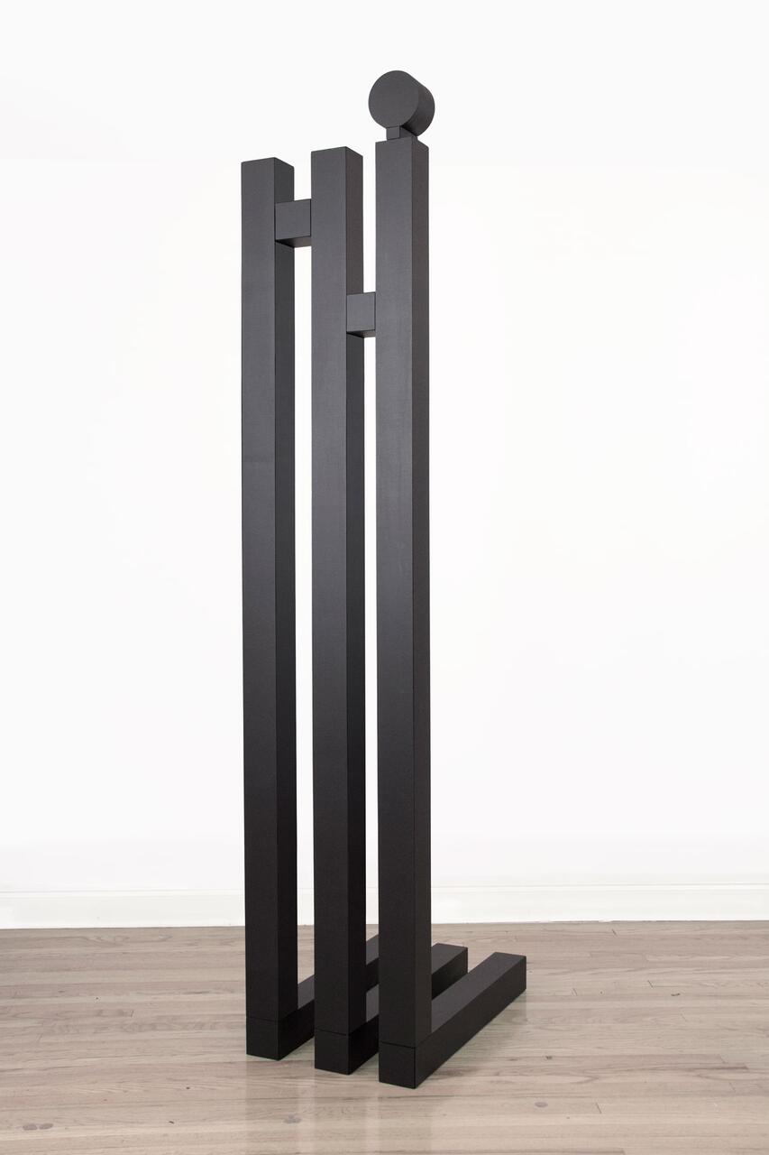 Tall, minimalist black sculpture with four vertical rectangular columns connected by horizontal beams, standing on a wooden floor against a plain white background.