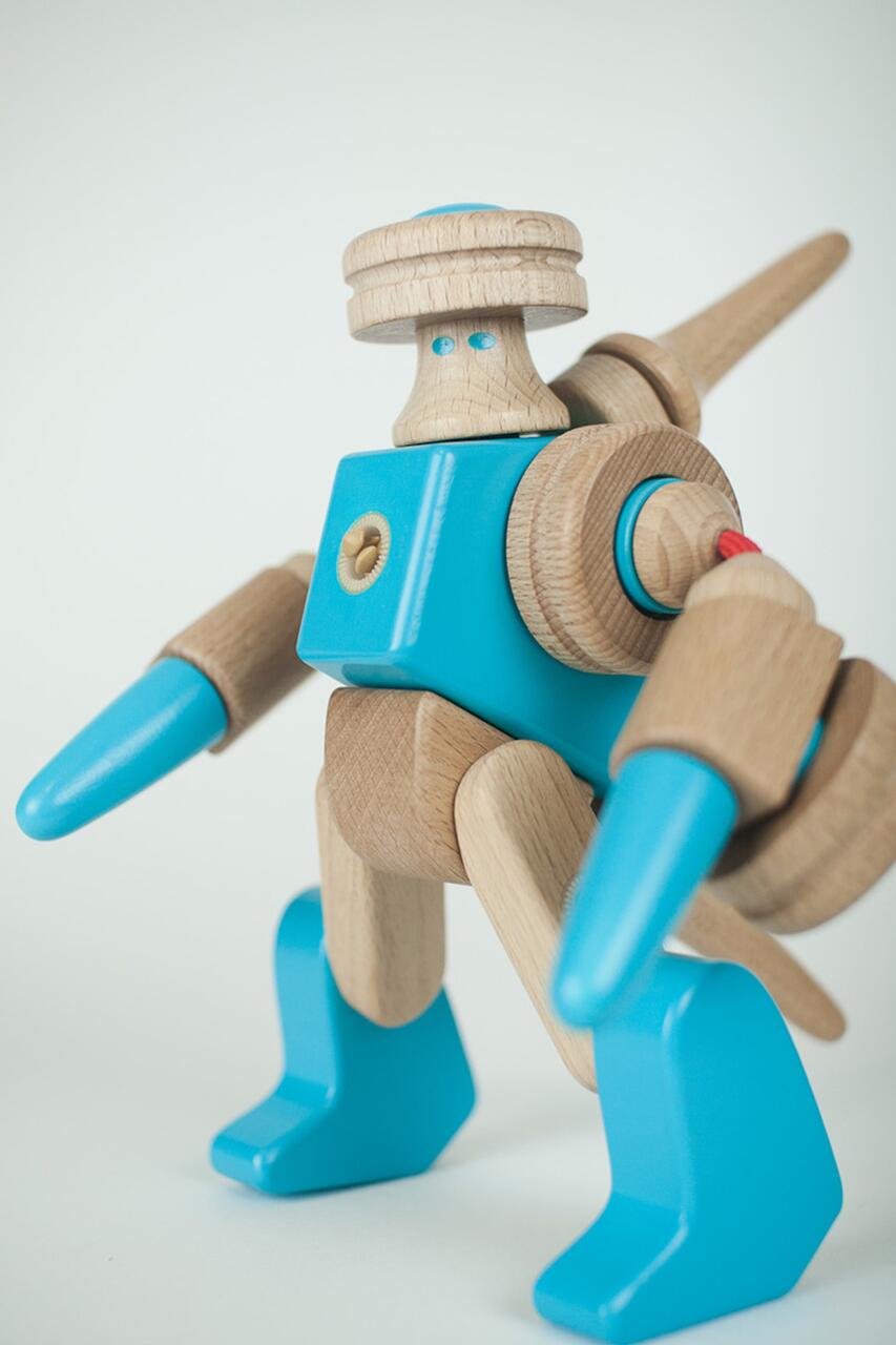 Wooden toy robot with a blue body and limbs, featuring cylindrical arms, rounded feet, and a small round head with blue details. The robot appears dynamic, standing on a white surface.