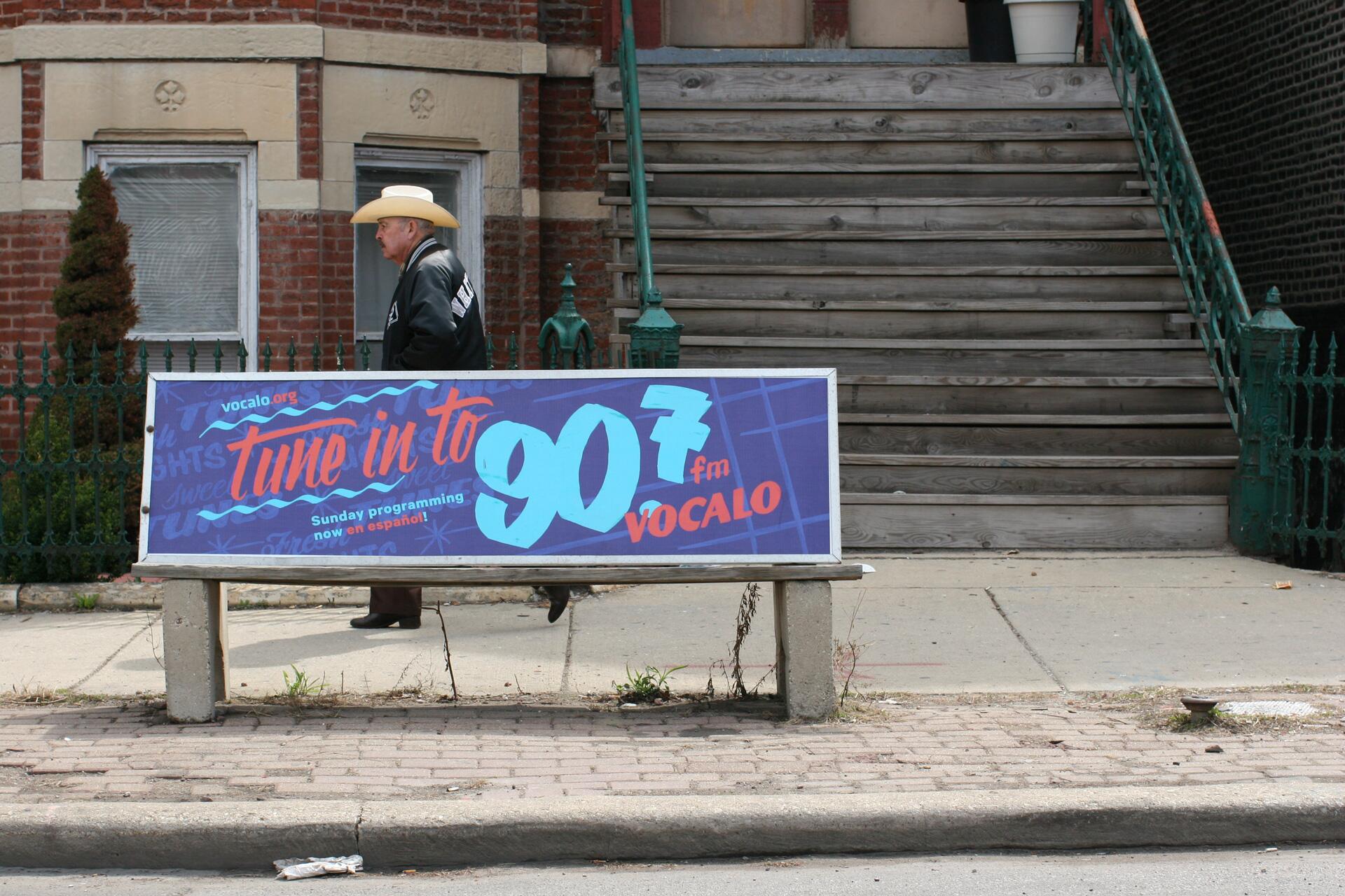 A man in a cowboy hat and jacket walks past a bench with an advertisement for Vocalo 90.7 FM in front of a brick building with wooden stairs.