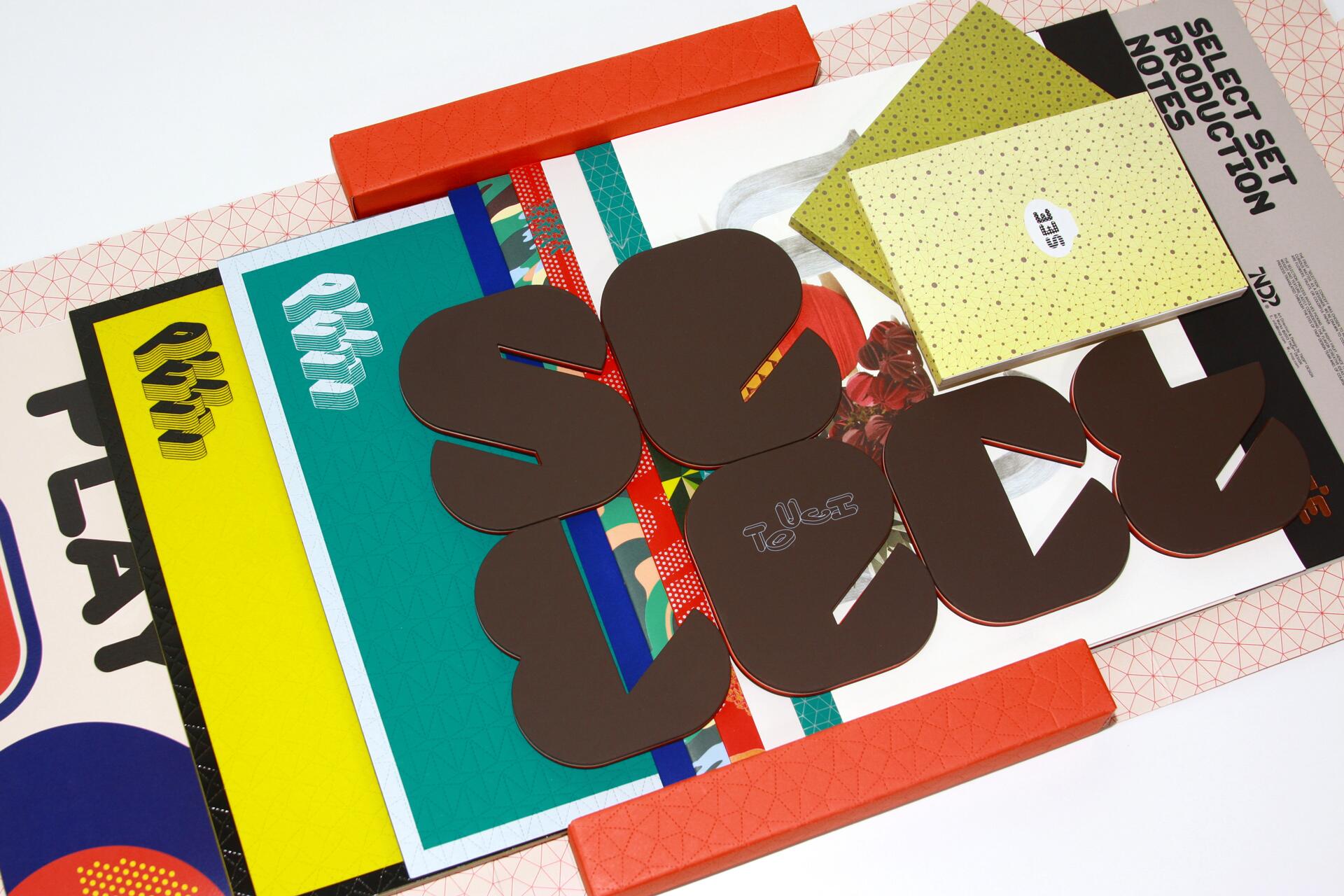 A collection of colorful printed materials and cut-out letters on a white background, with orange geometric borders. Various textures, colors, and patterns are visible, including teal, yellow, and dotted designs.