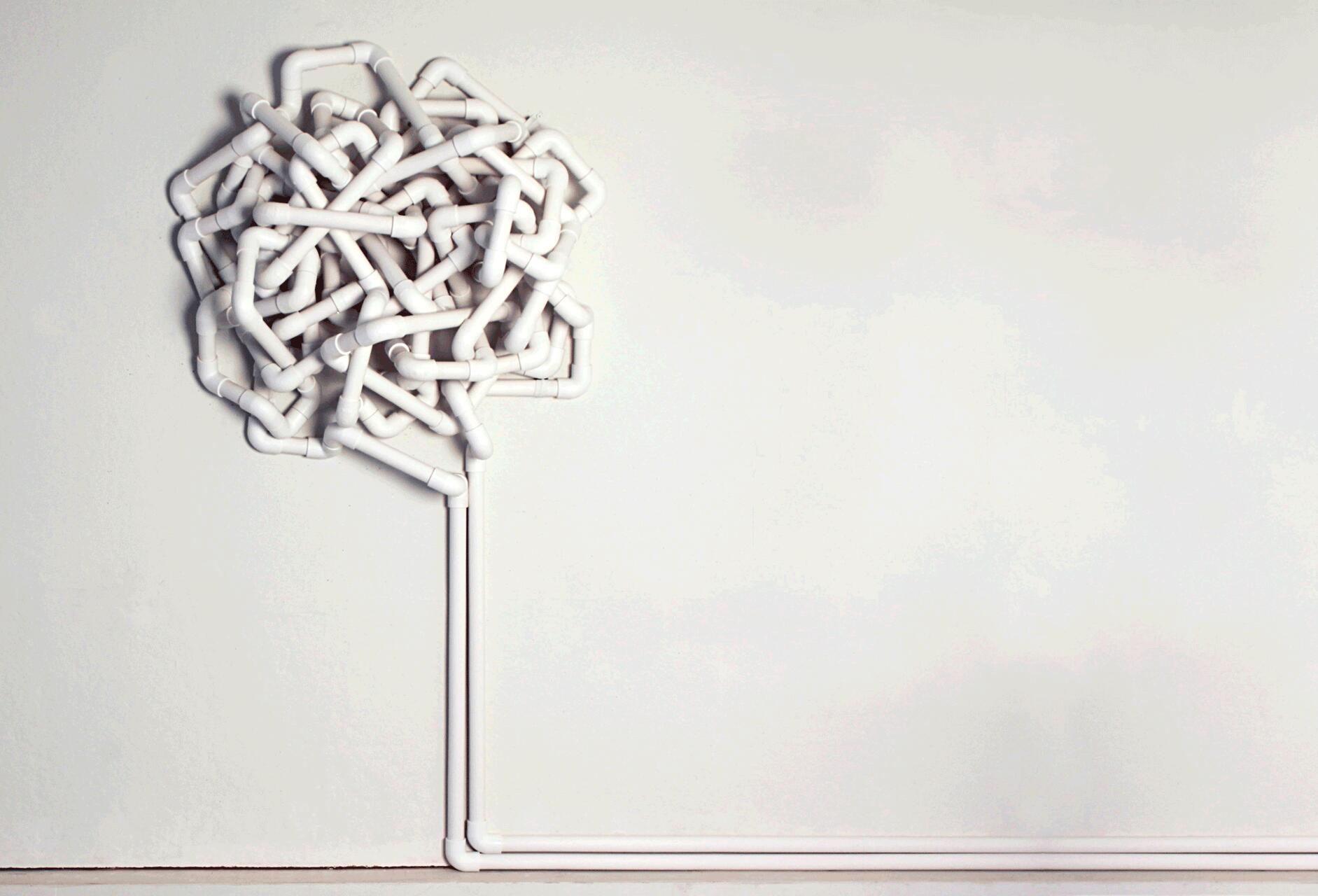 White PVC pipes arranged in a complex, tangled pattern resembling a tree against a plain white wall.