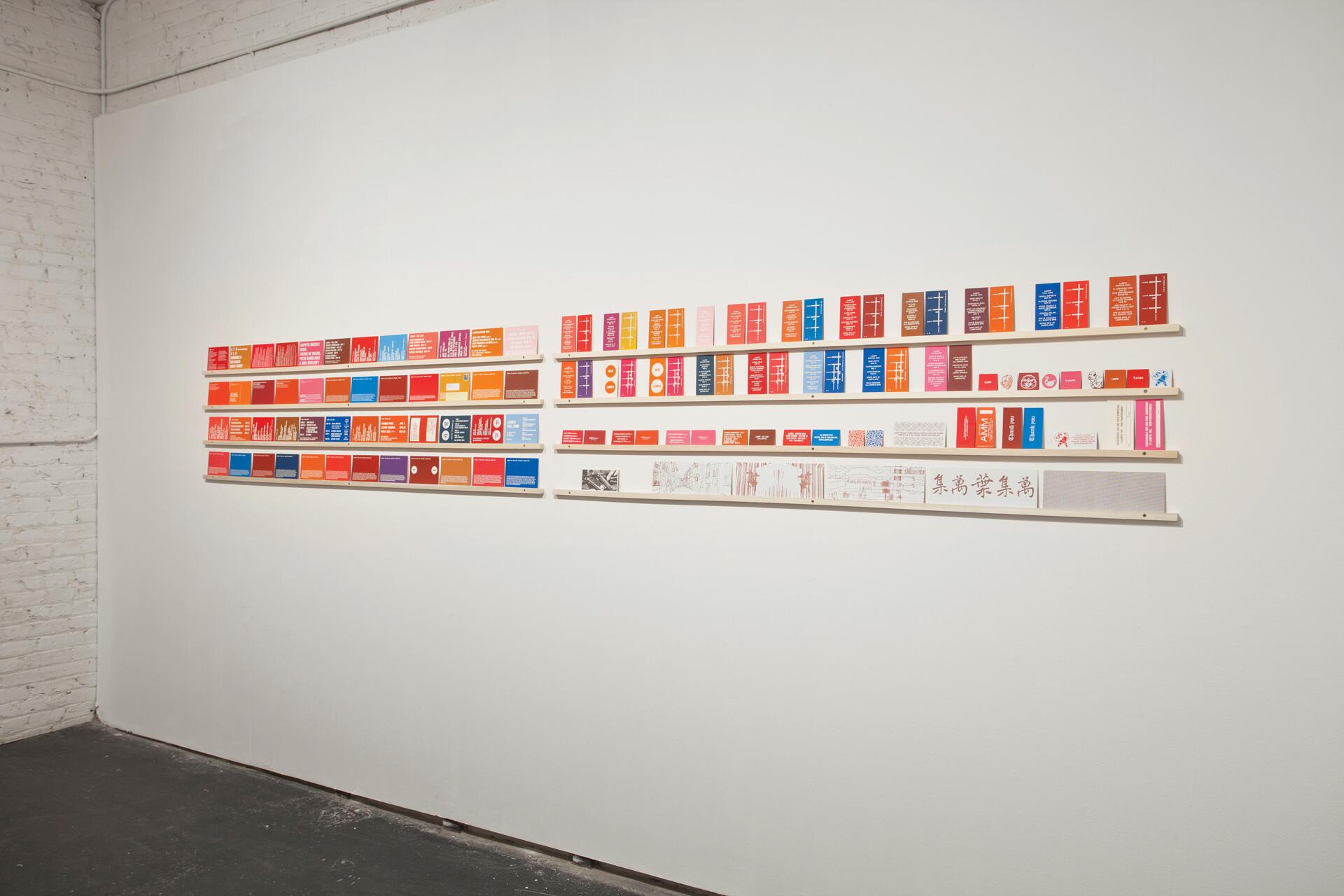 Colorful exhibition displays rows of variously colored cards with text, mounted on a white gallery wall. The cards are arranged in horizontal lines, showcasing a range of bright hues like red, blue, orange, and pink.