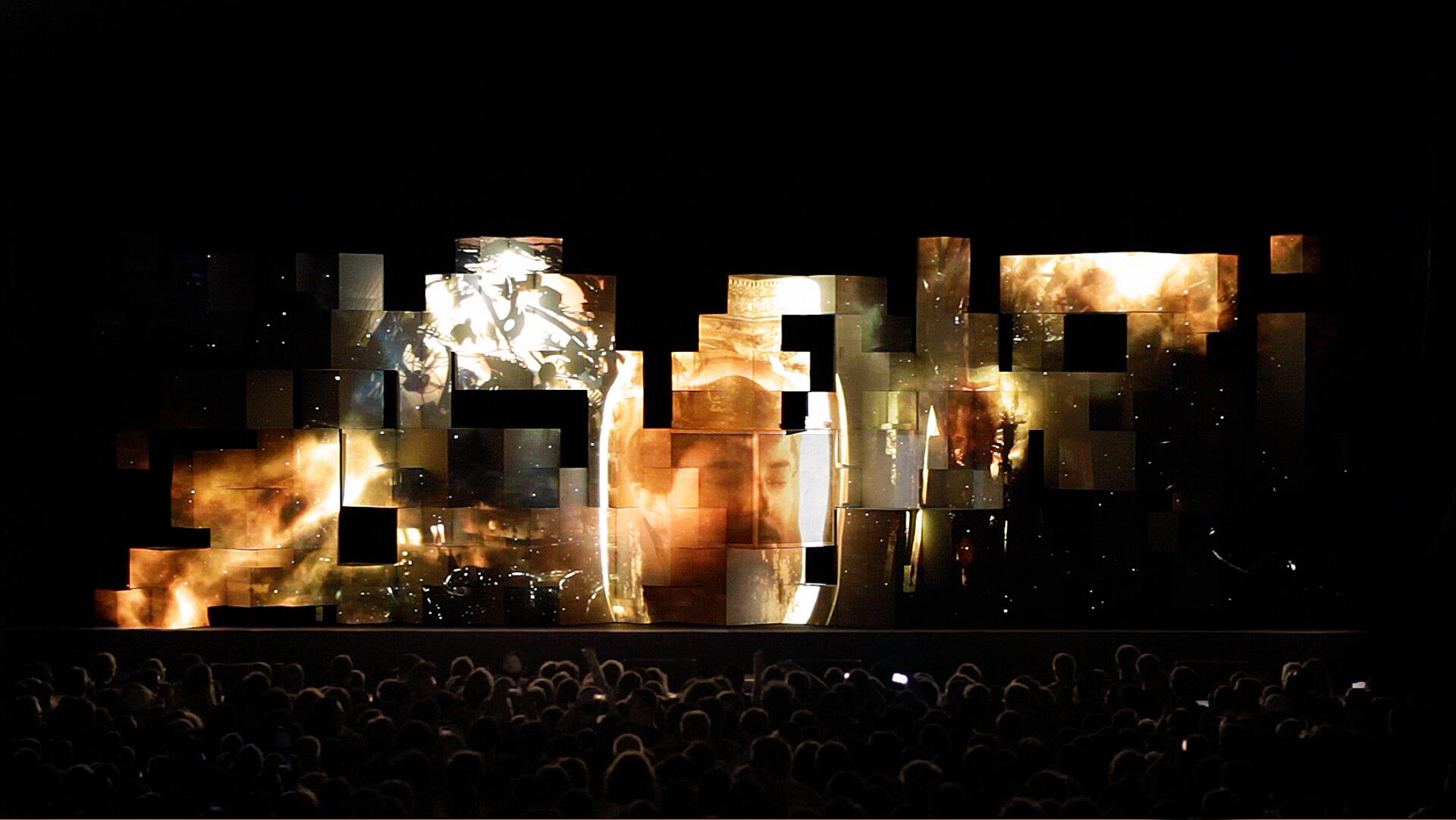 Abstract digital art installation with glowing geometric shapes and projections of faces and lighting effects against a dark background, observed by a crowd.