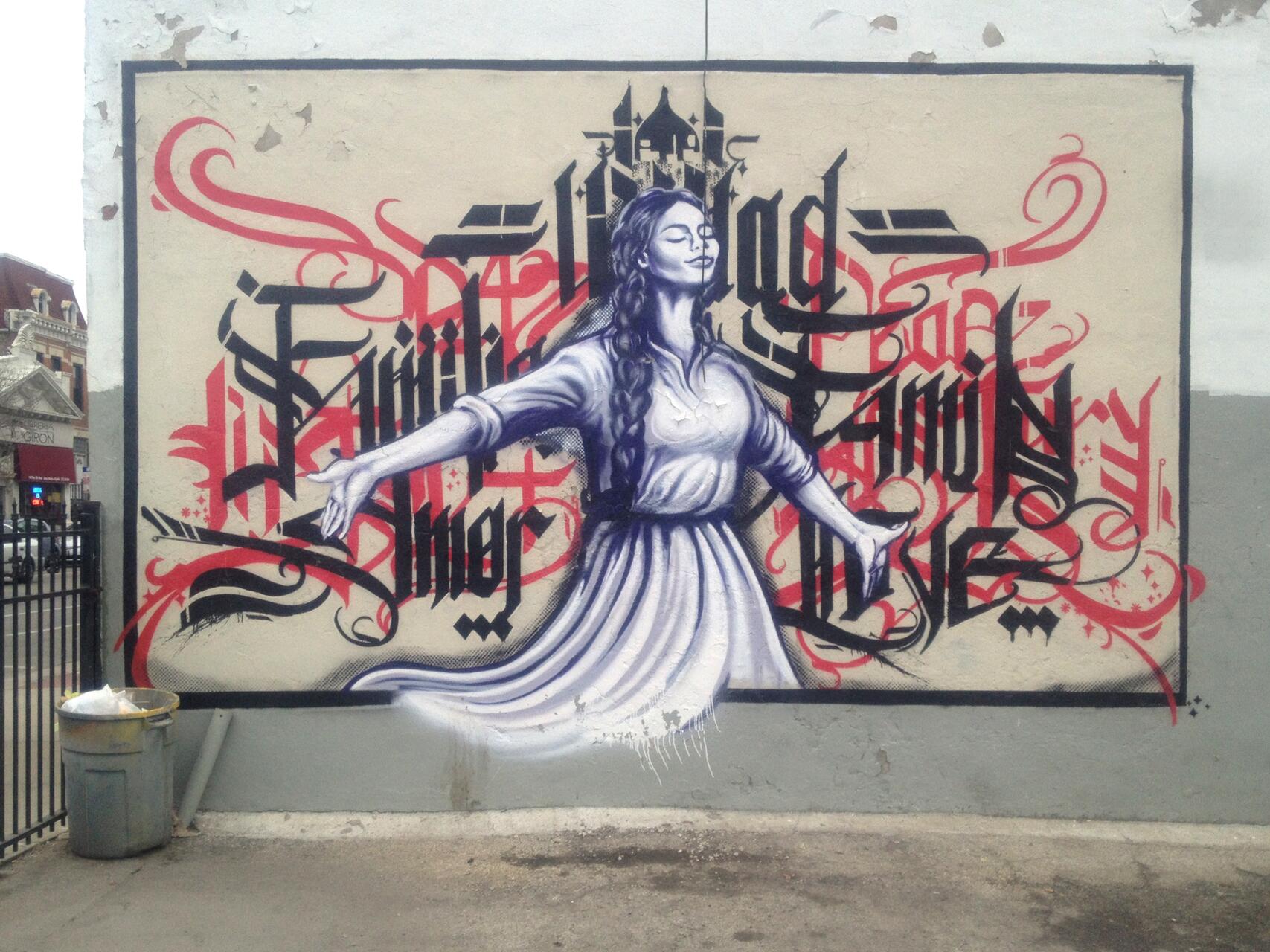 A mural on a wall depicts a woman with outstretched arms, eyes closed in a peaceful expression. She is set against intricate black and red calligraphic text. The background is a worn, weathered wall with chipping paint.