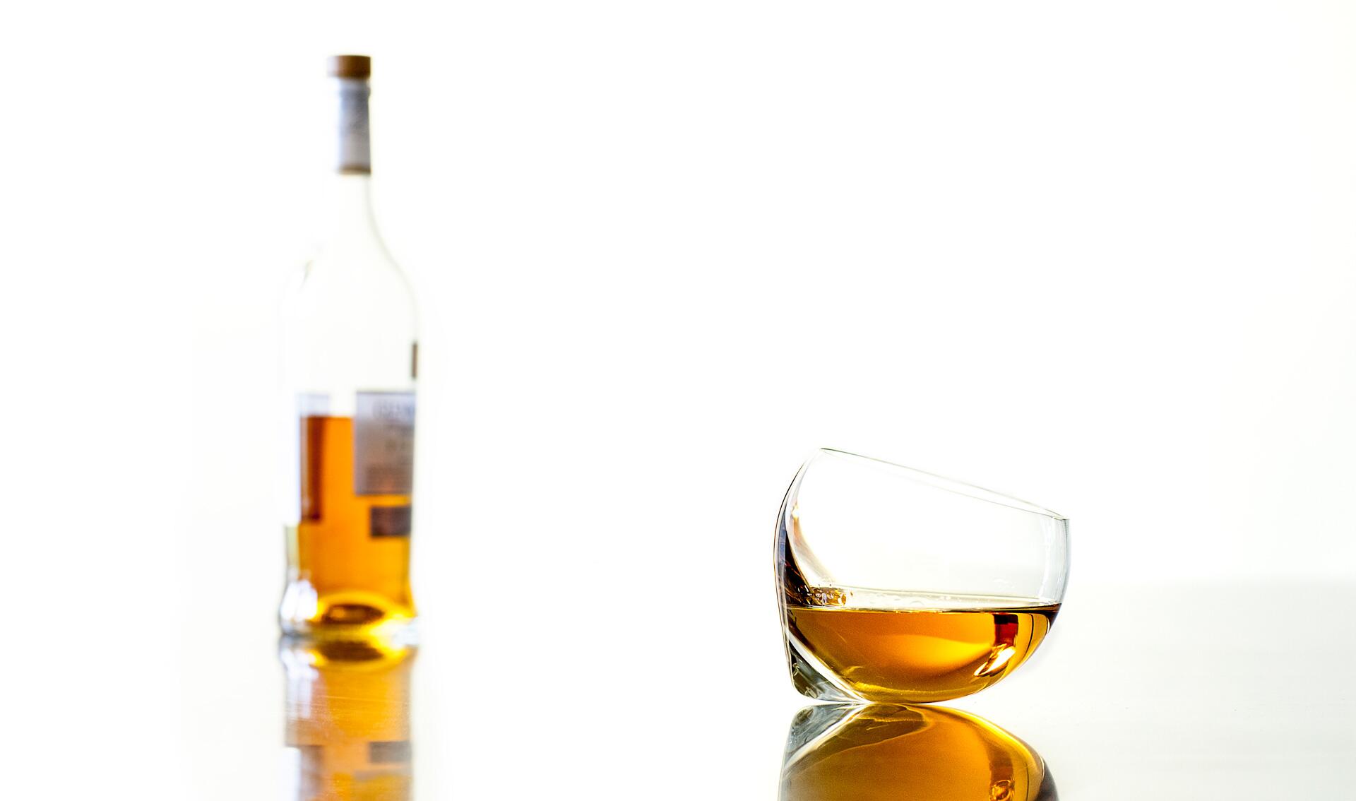 A tilted glass with amber liquid reflecting on a glossy surface, with a blurred bottle of similar liquid in the background against a white backdrop.