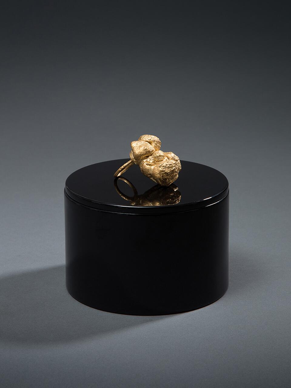 Gold ring with a textured, abstract design sits on a glossy black cylinder against a gray background.