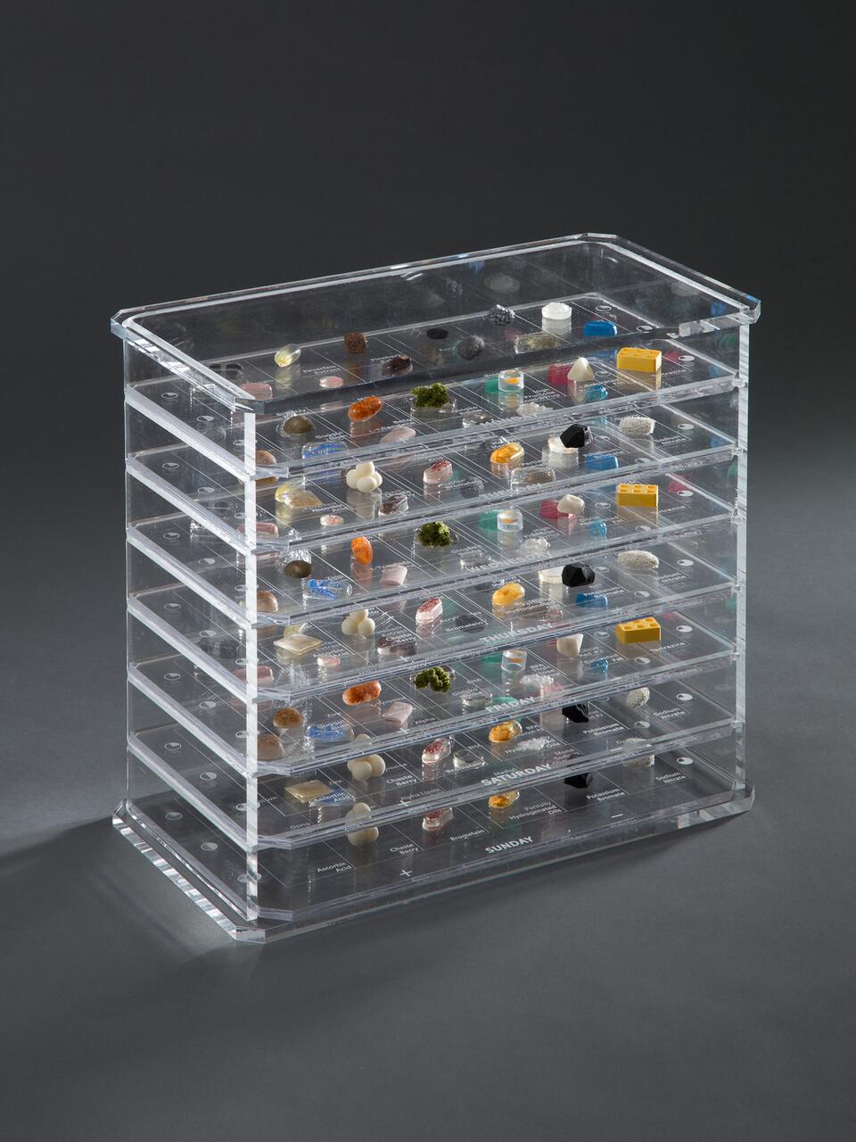 Clear acrylic case with five horizontal drawers, each containing various small objects like pills, stones, and colorful items, organized separately.