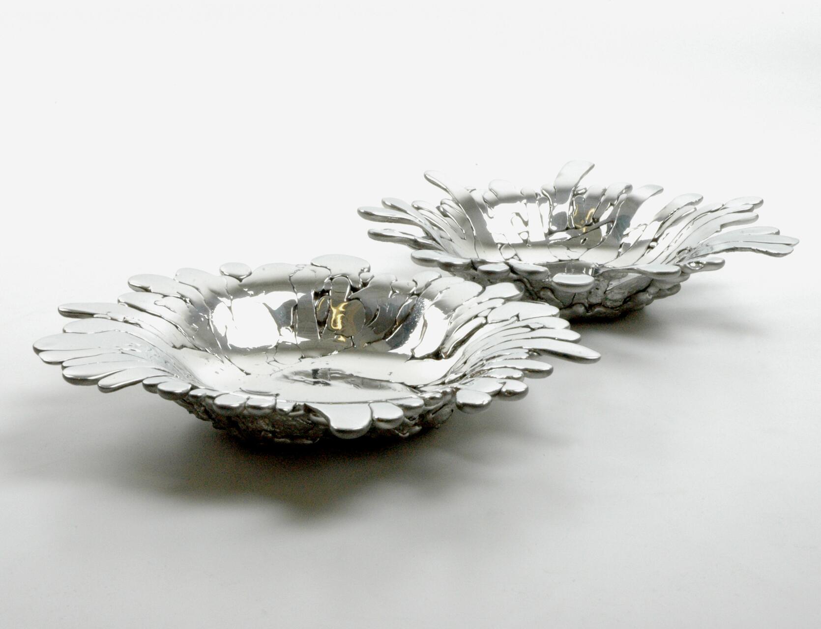 Two sculptural silver bowls with petal-like edges, resembling flowers, on a white background. They have a reflective, metallic finish and are positioned diagonally.