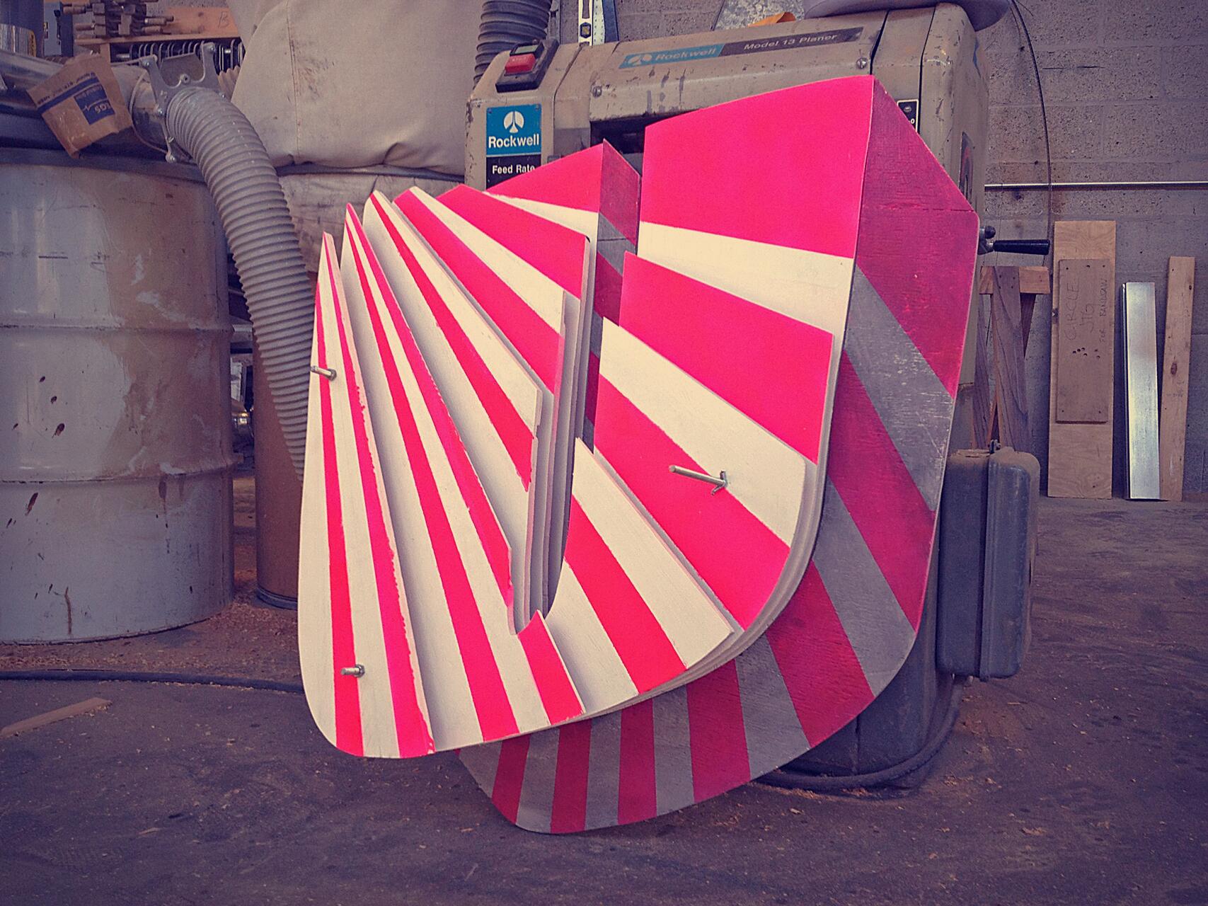 Red and white striped geometric sculpture leaning against industrial machinery in a workshop, surrounded by wood planks and metal pipes, under warm lighting.