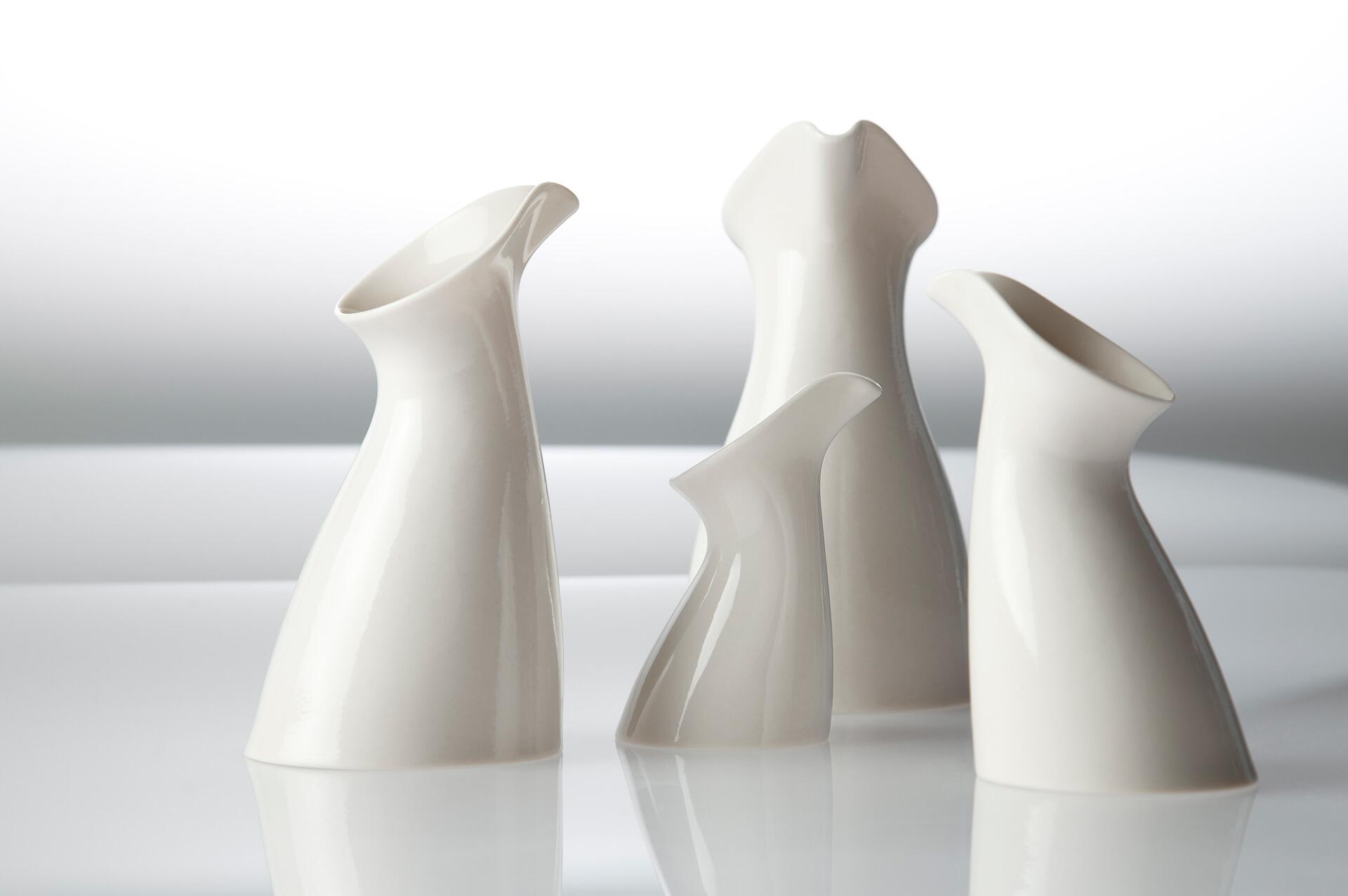 Four white, modern ceramic pitchers with curved forms are arranged on a reflective surface against a gradient background.