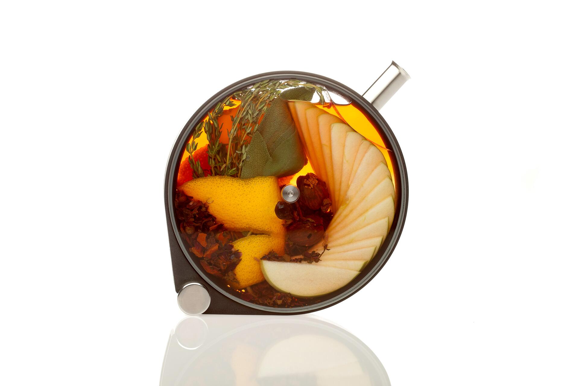 Top view of a glass teapot filled with orange-hued tea, floating apple slices, citrus peels, herbs, and spices, against a white background.