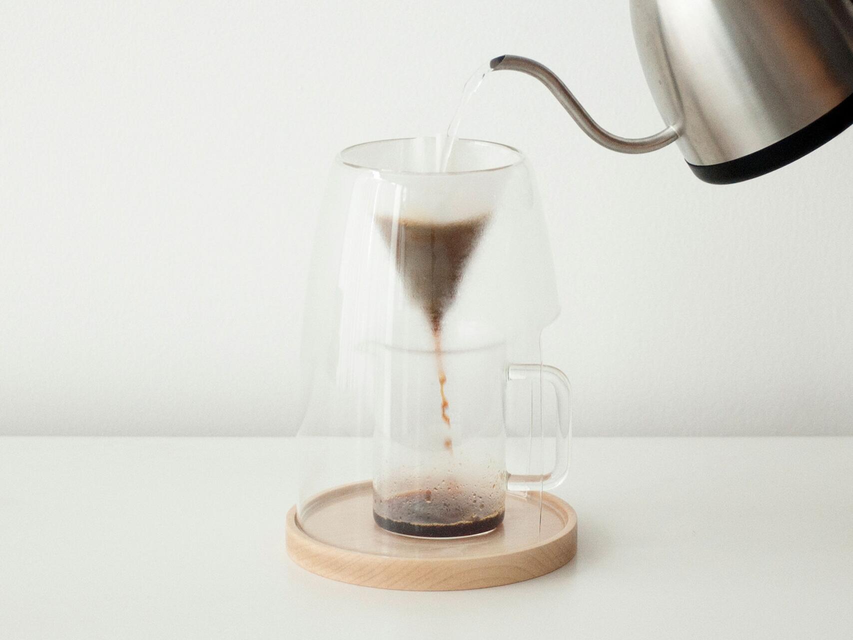 Pour-over coffee being made, with hot water from a stainless steel kettle flowing into a clear glass dripper set on a wooden tray.
