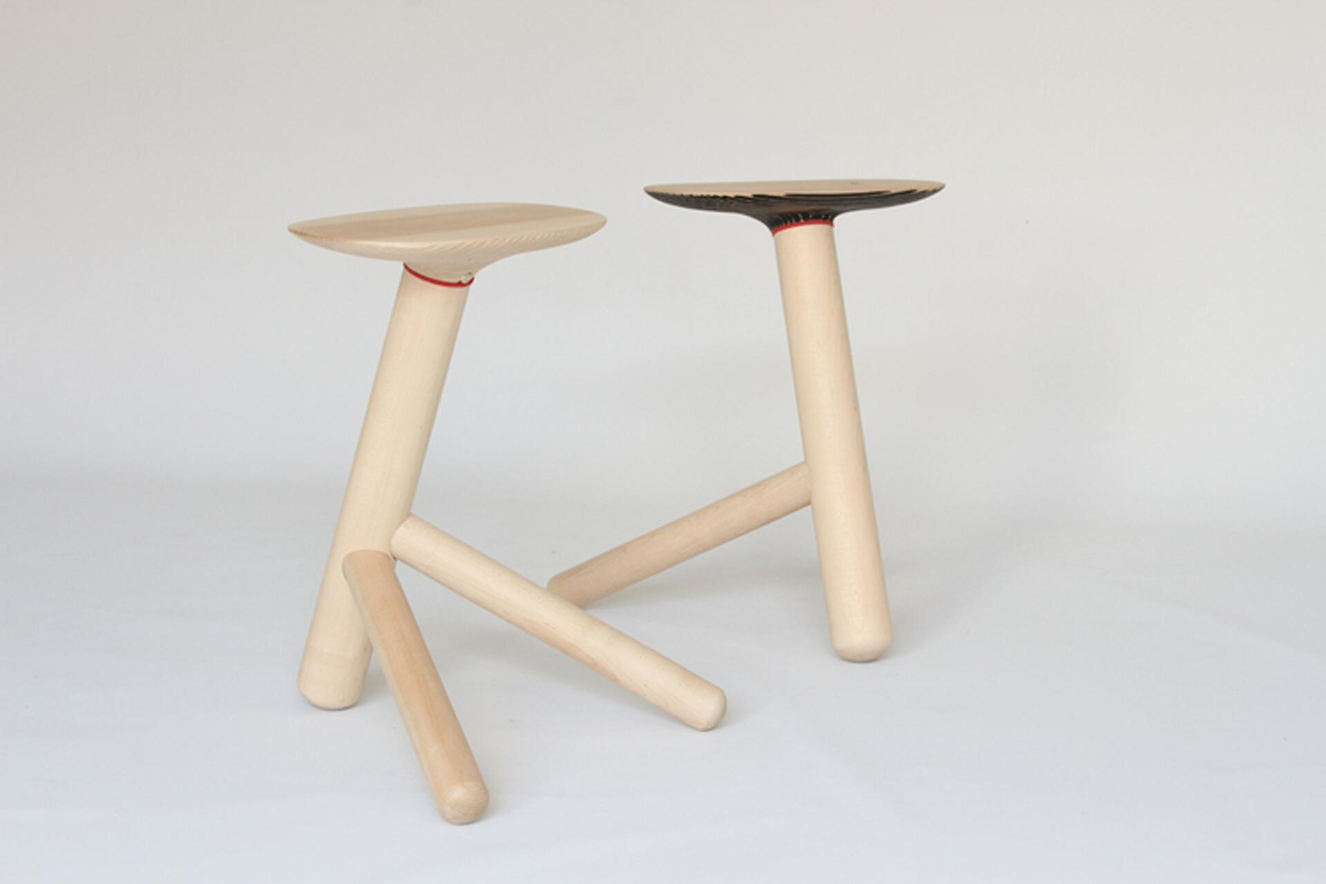 Two minimalist, three-legged wooden stools with angled legs on a white background. One has a natural finish, and the other has a dark seat with red accents.