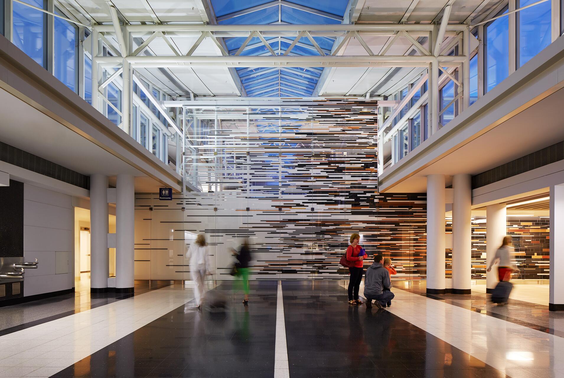 Spacious modern hallway with high glass ceiling, abstract wall art of horizontal lines in various colors. People walking and interacting, one person kneeling with a child, others carrying luggage. Bright lighting creates a clean and open atmosphere.