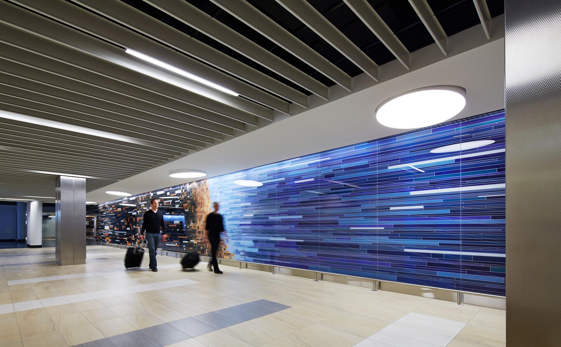 Two people with suitcases walk through a modern hallway with a colorful, abstract wall art featuring blue and purple horizontal lines. Bright ceiling lights illuminate the scene.