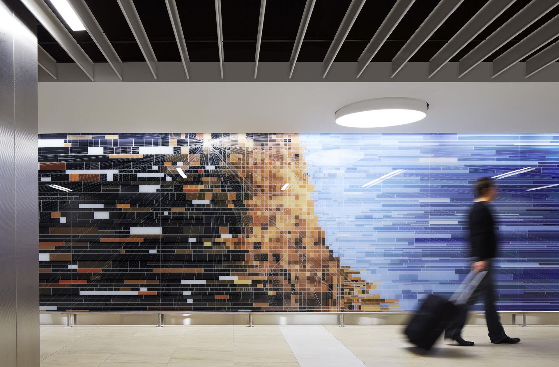A person walks with a suitcase past a large abstract mural with pixelated patterns in shades of black, brown, and blue. The setting appears modern, with a sleek ceiling and bright lighting.