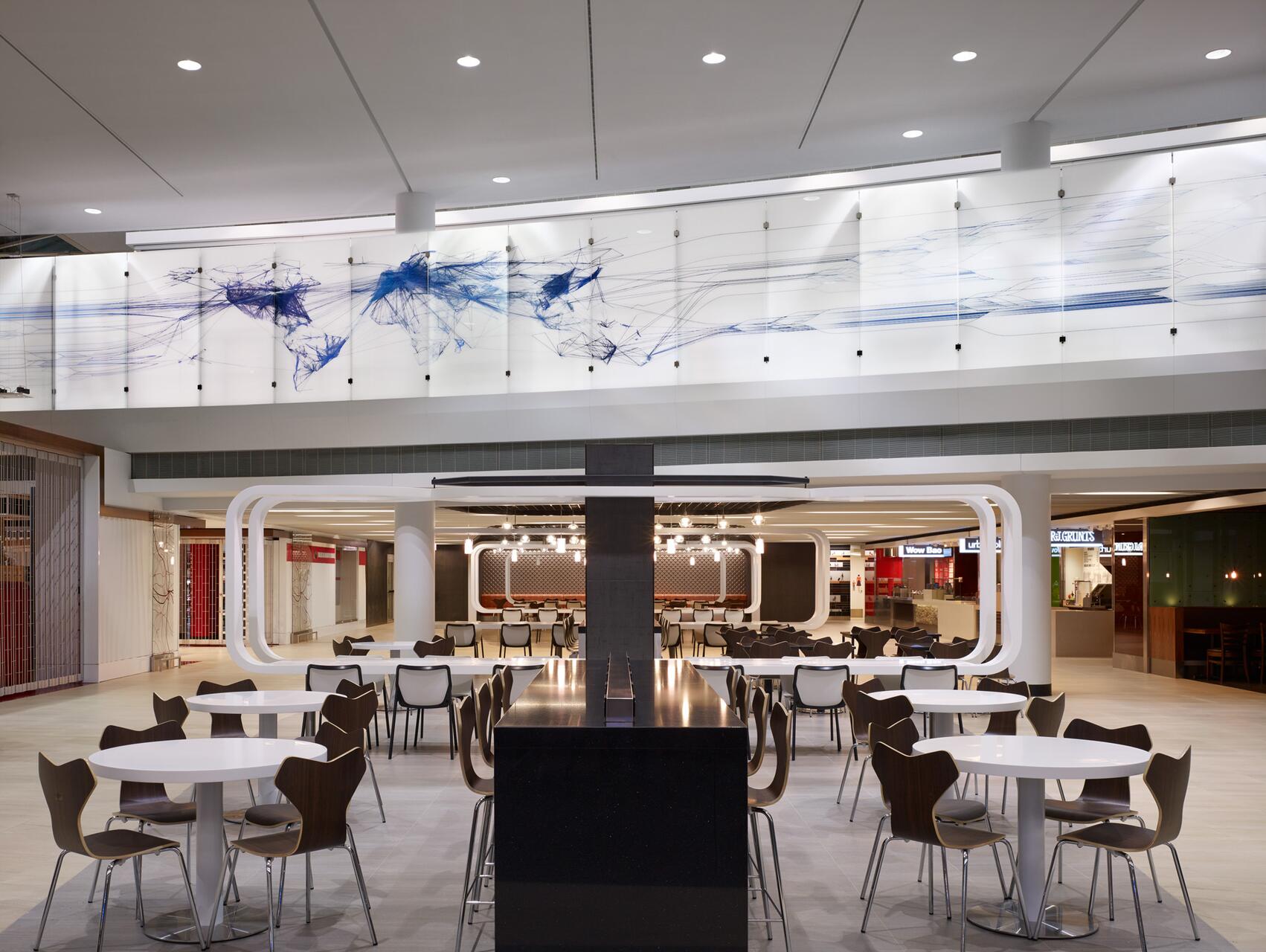 Modern food court with white and wood chairs and round tables, under a high ceiling with bright lighting. A decorative wall with abstract blue line art map is visible above, creating a spacious, contemporary atmosphere.