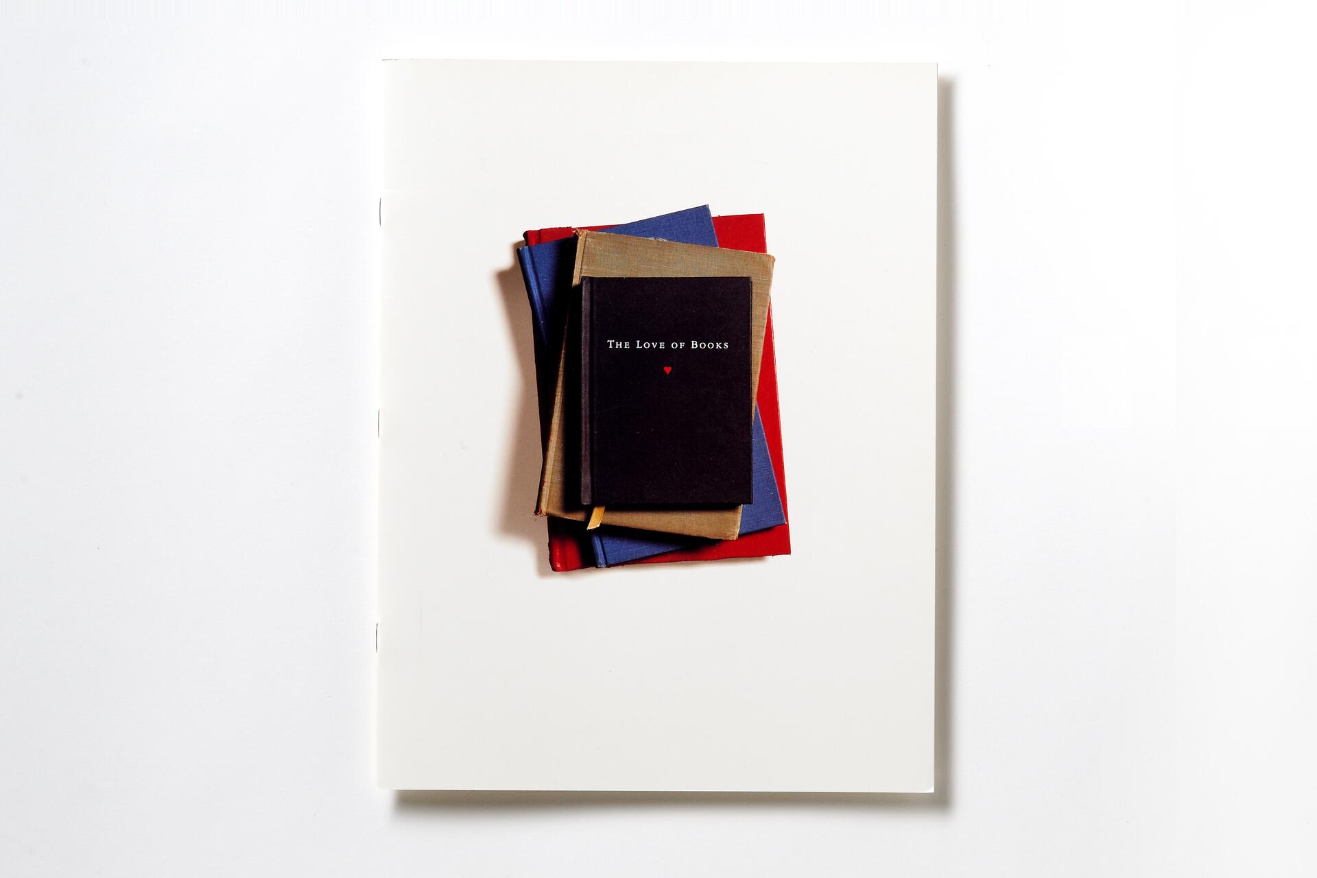 A stack of books with red, blue, and brown covers on a plain white background. The top book is black with the title "The Love of Books" in white lettering and a small red heart below.