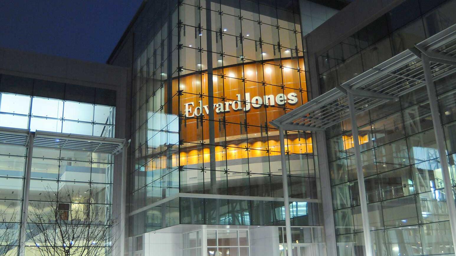Modern glass building at night, with illuminated letters spelling "Edward Jones" on an interior wooden panel, reflecting warm light.
