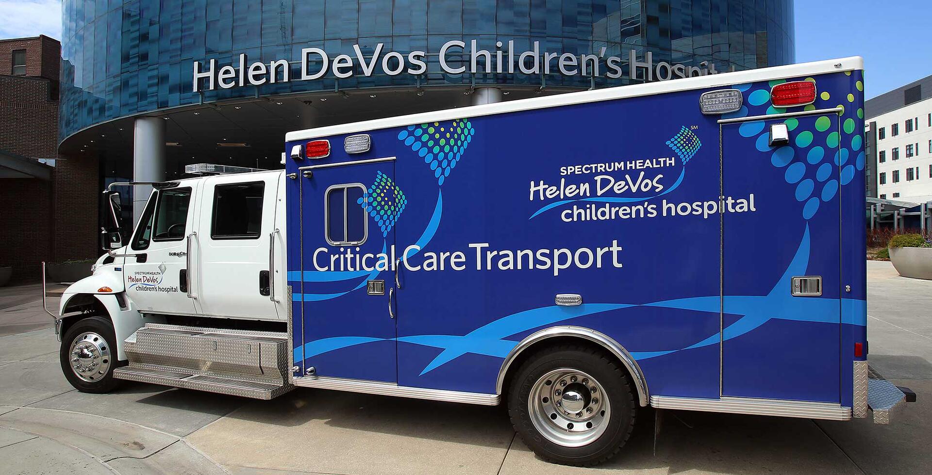 Blue and white ambulance parked in front of Helen DeVos Children's Hospital, with "Critical Care Transport" and colorful logos on its side.