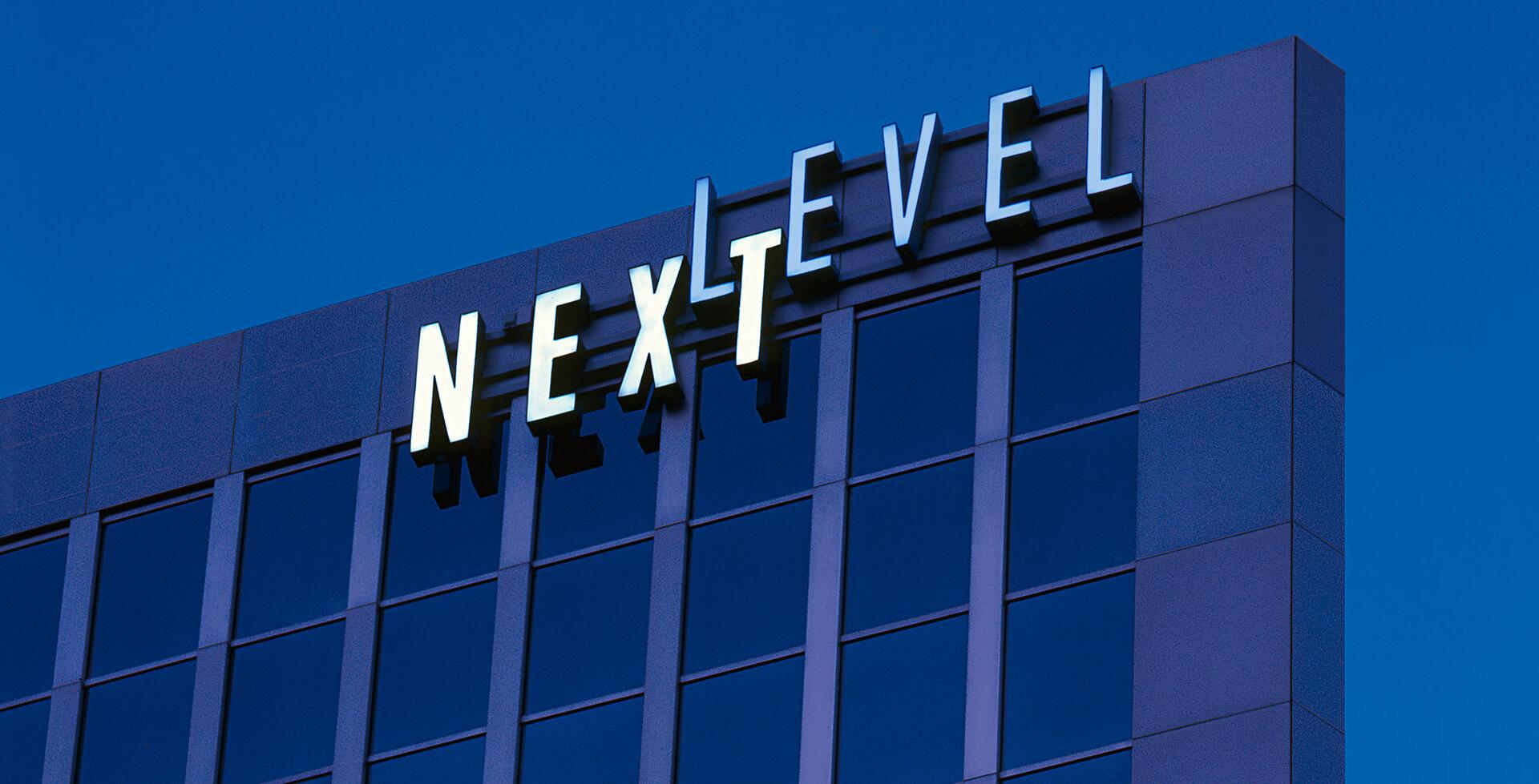 Modern office building facade with reflective blue windows and large illuminated sign reading "NEXT LEVEL" at the top.