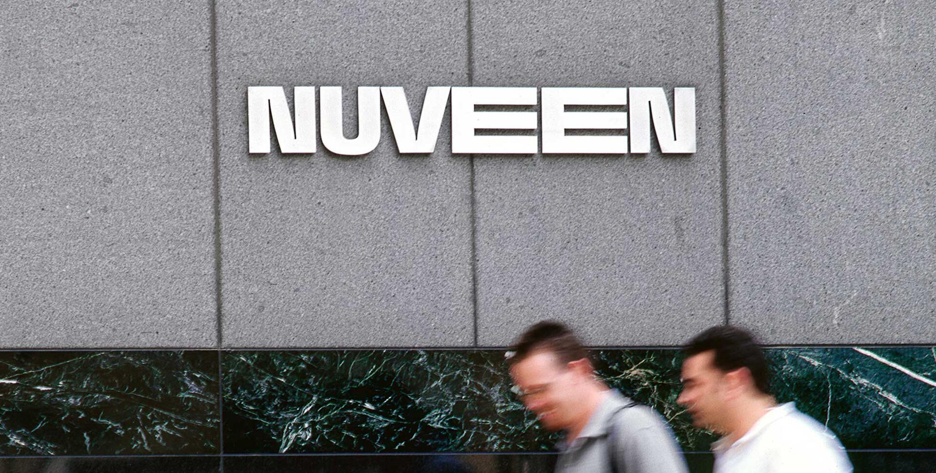 Blurred pedestrians walk past a building with "NUVEEN" in large white letters on a gray stone wall, with a dark green marble strip below.