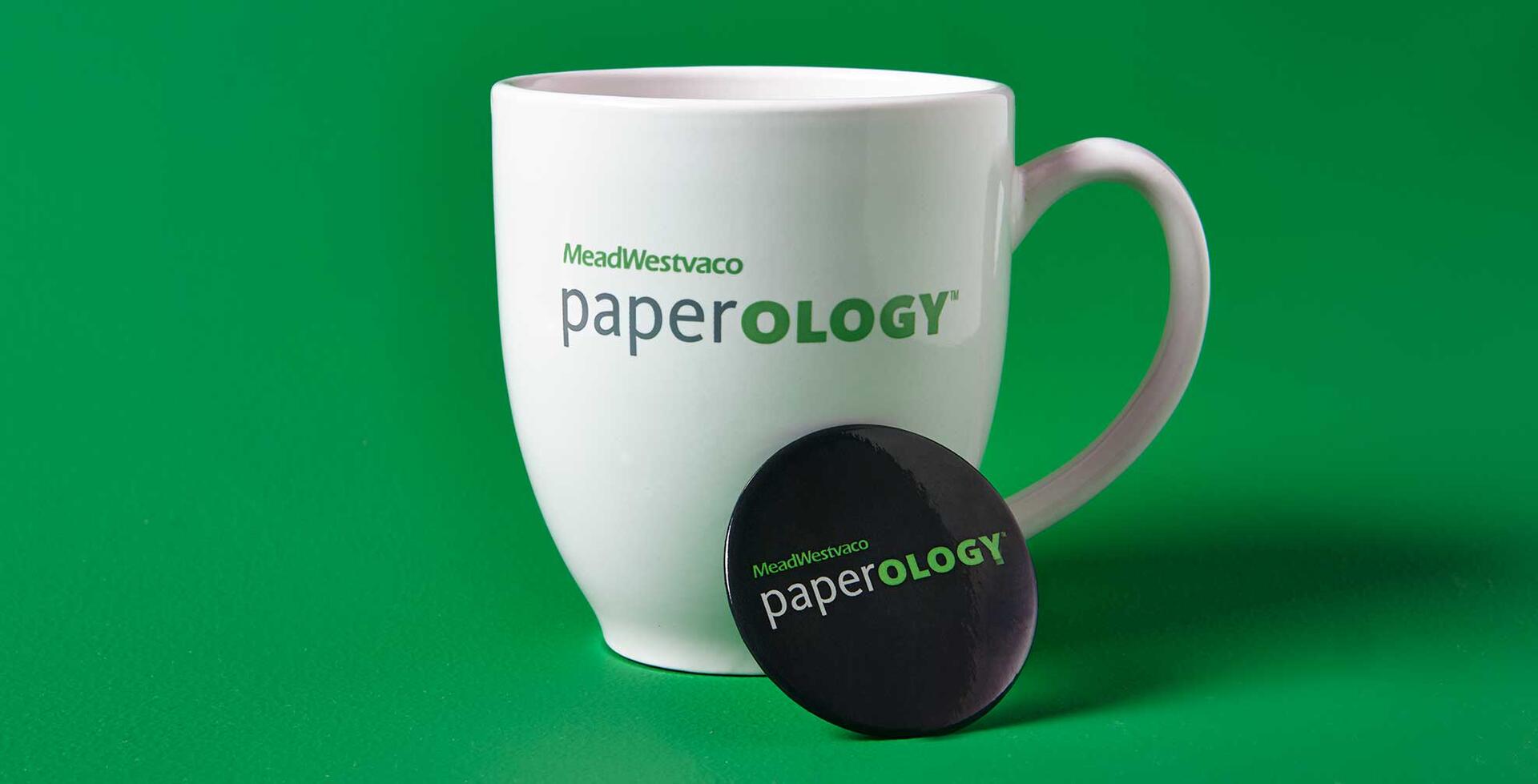 White mug with "MeadWestvaco paperOLOGY" logo in green and gray, placed on a green background. A round black pin with the same logo leans against it.