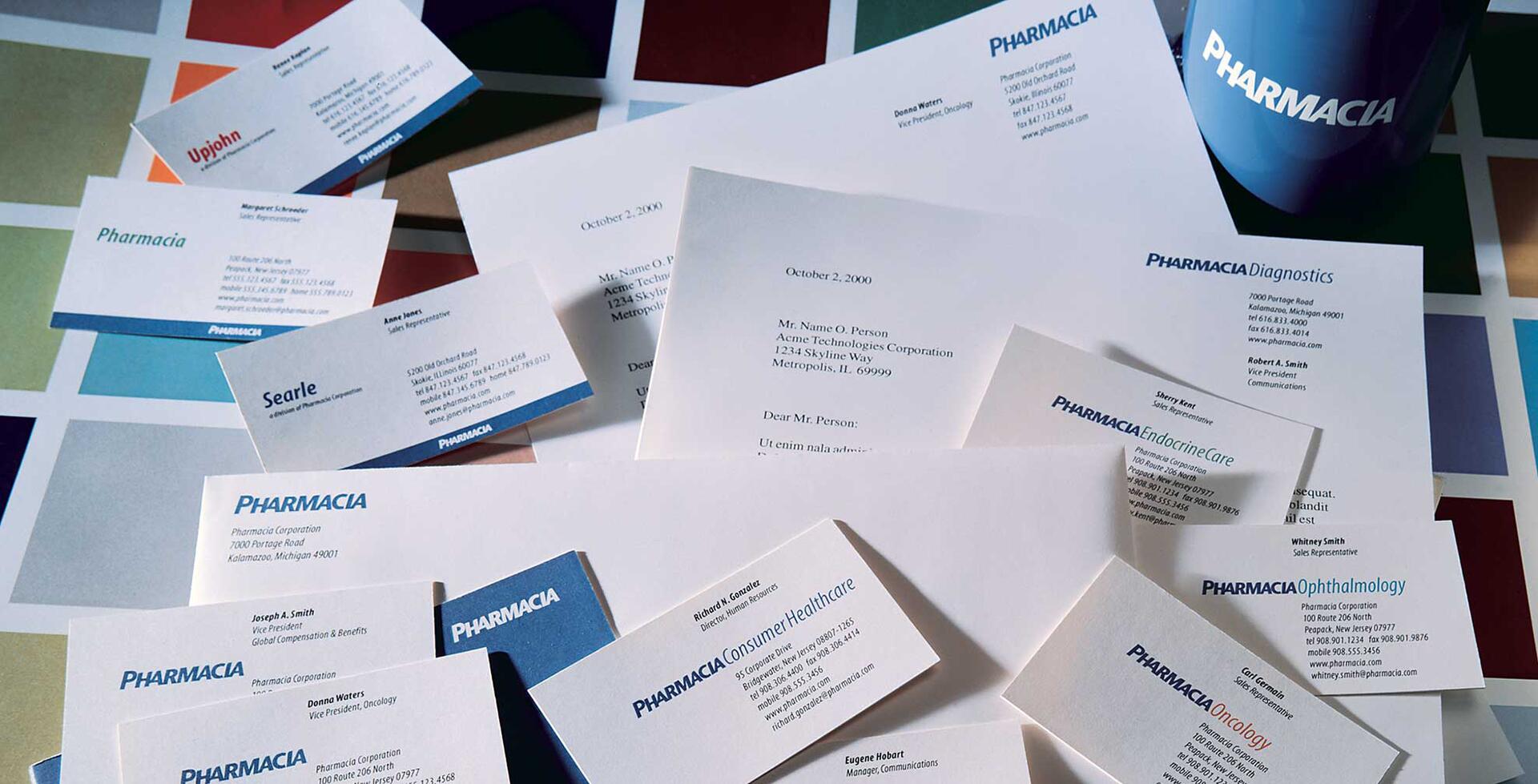 Business cards and letters from Pharmacia Corporation, arranged on a colorful, tiled surface. A blue mug with "PHARMACIA" logo is visible, surrounded by cards bearing various department names and individuals.