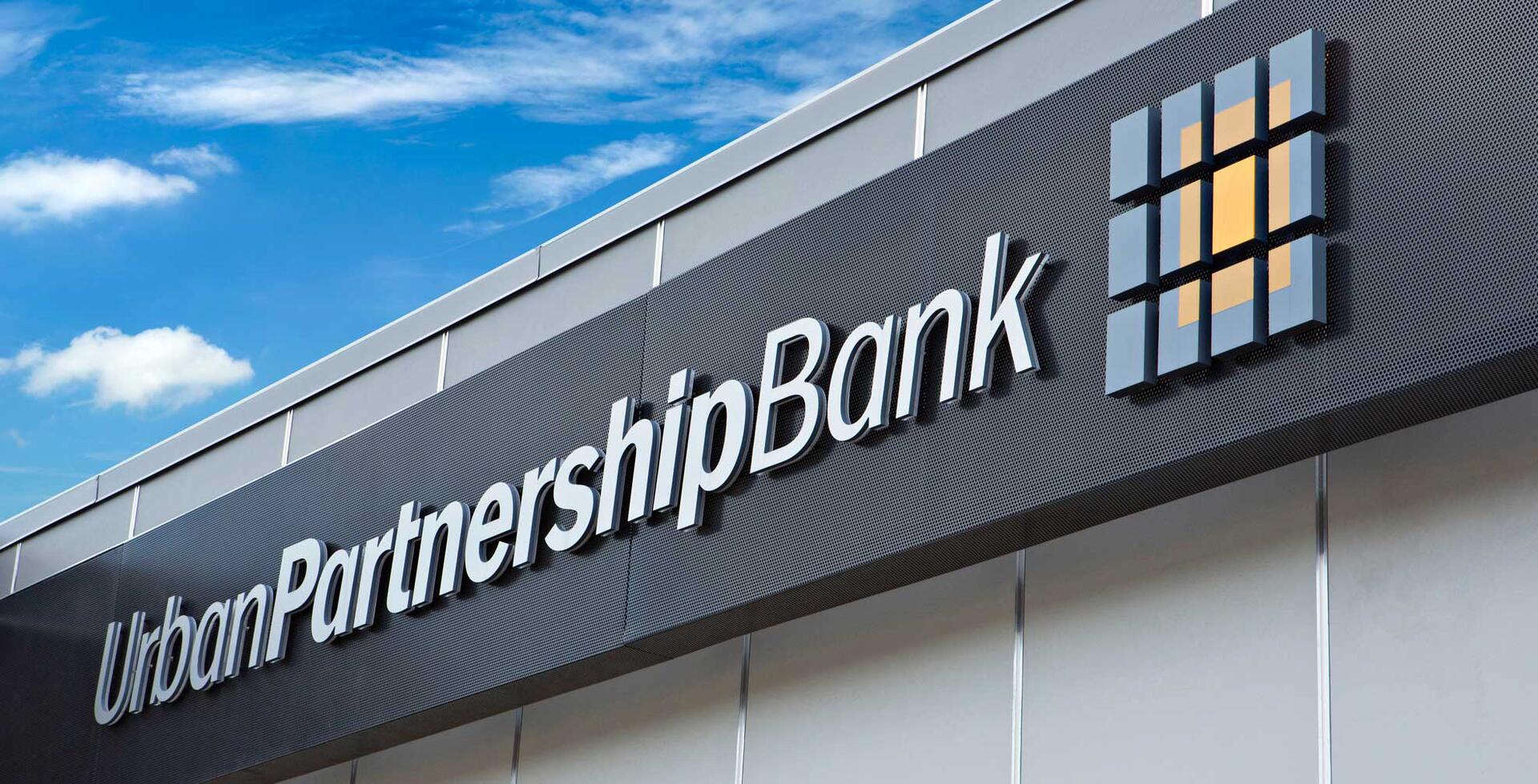 Building facade with "Urban Partnership Bank" signage and a grid-like logo under a blue sky with clouds.