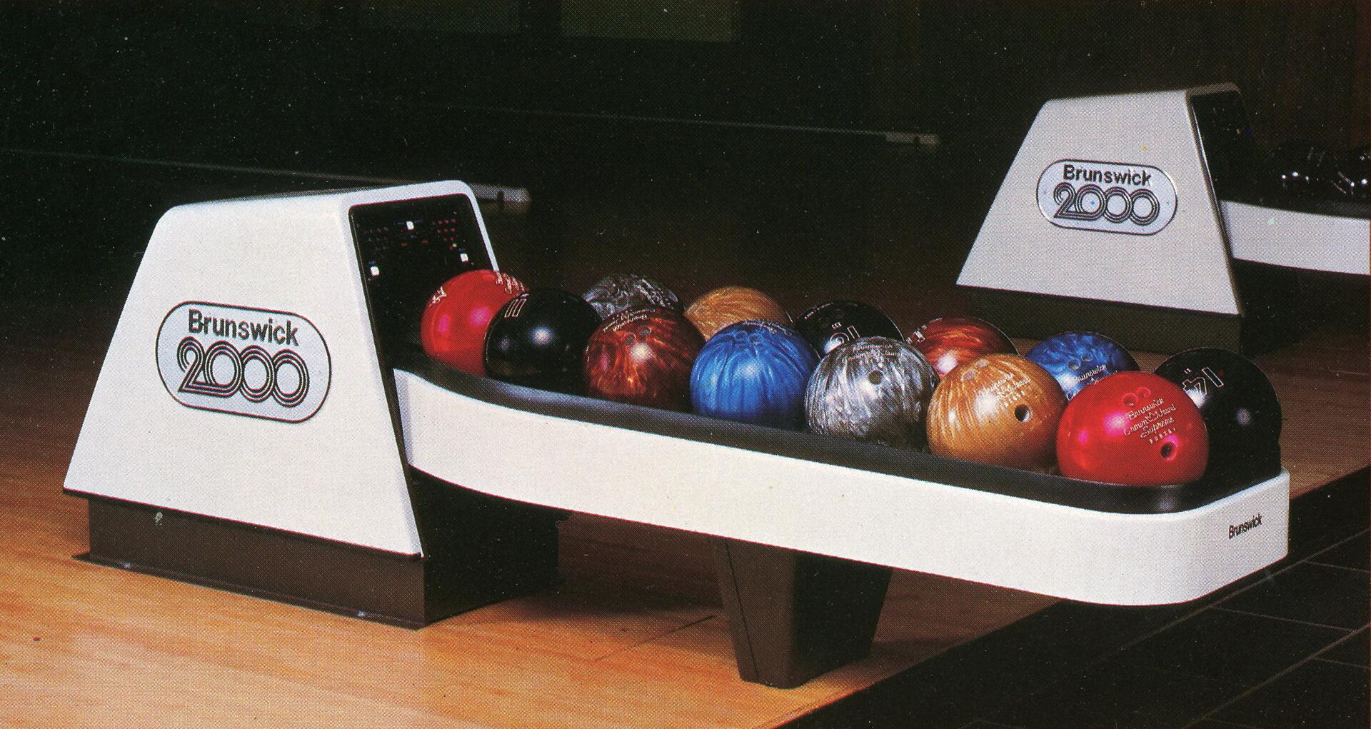 Bowling ball return system labeled "Brunswick 2000" on a wooden floor, holding an array of colorful bowling balls including red, blue, black, and gold.