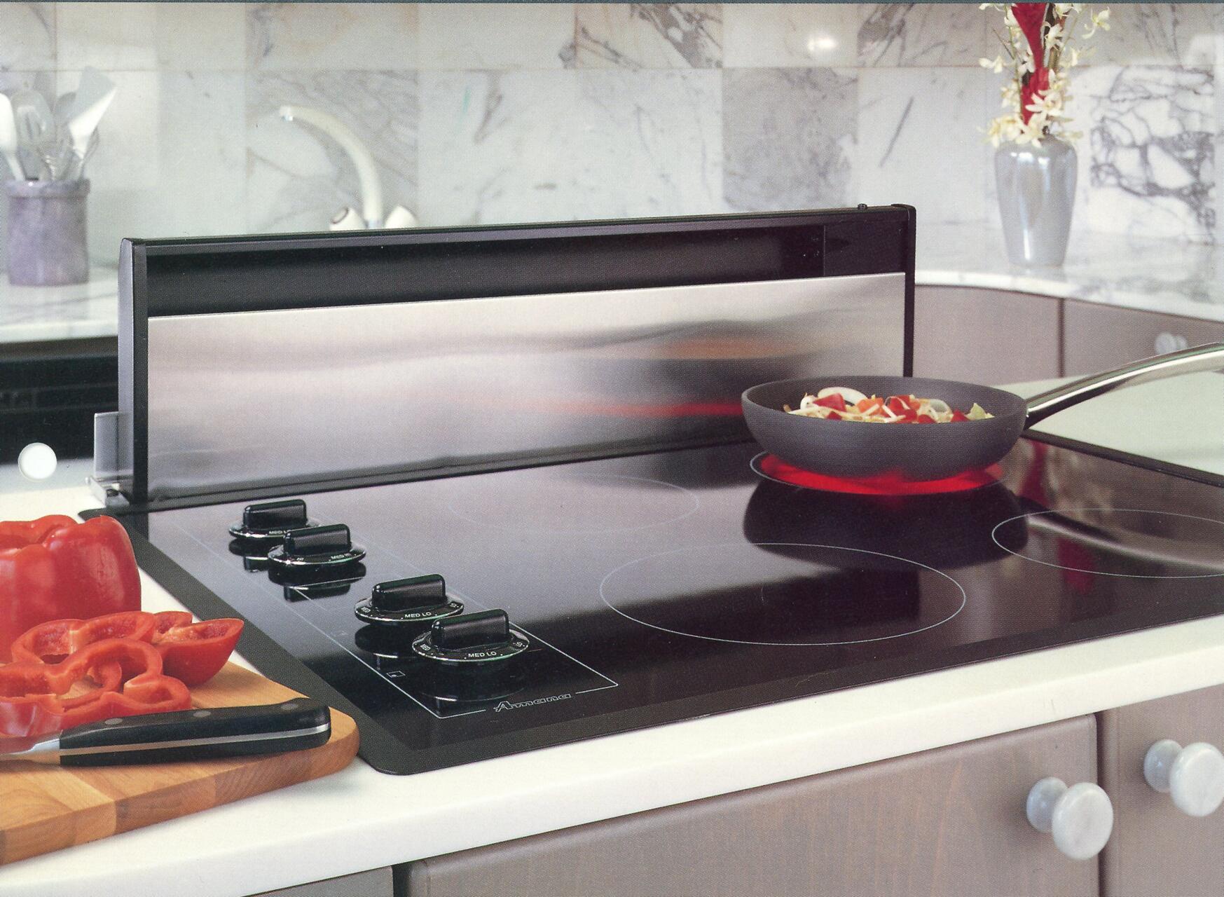 Modern kitchen with an electric stovetop. A black pan with vegetables is on a lit burner. Sliced red bell peppers are on a wooden cutting board with a knife. Marble backsplash and a vase with flowers in the background.
