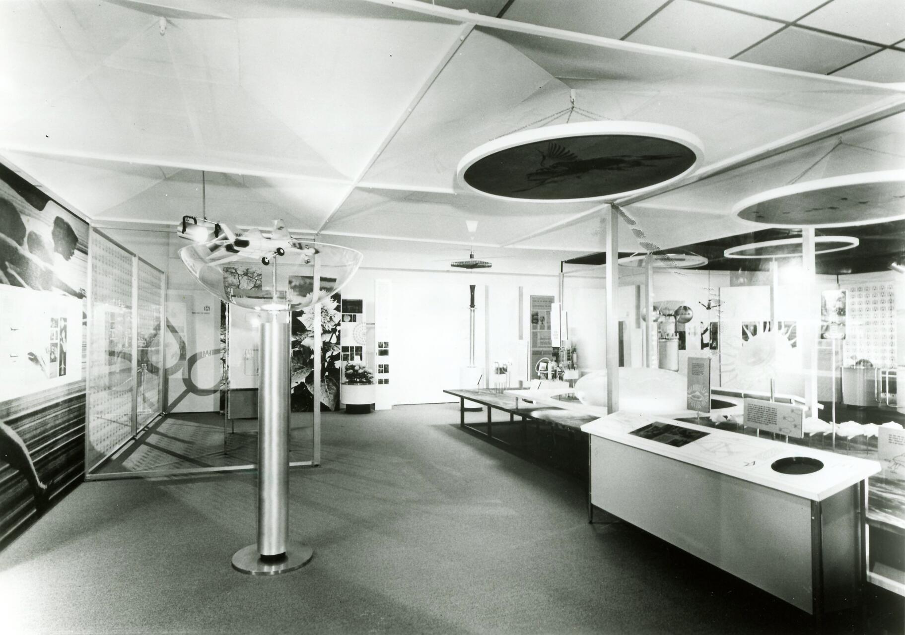 Black and white photo of a modern exhibition room with displays on aerodynamics. Circular models hang from the ceiling, and a variety of educational panels and diagrams are on tables and walls.
