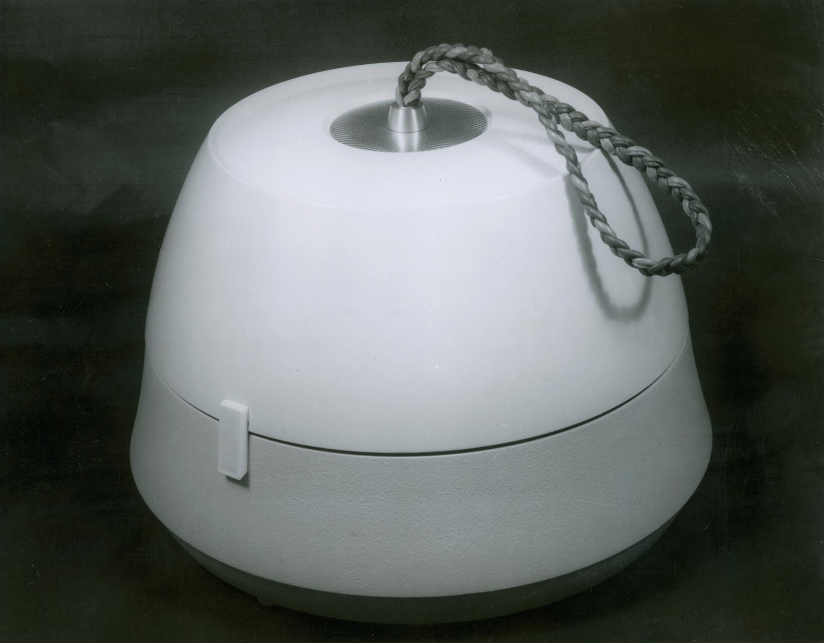 A round, smooth object with a rope handle on top, set against a dark background. The surface appears glossy with a small latch on the side.