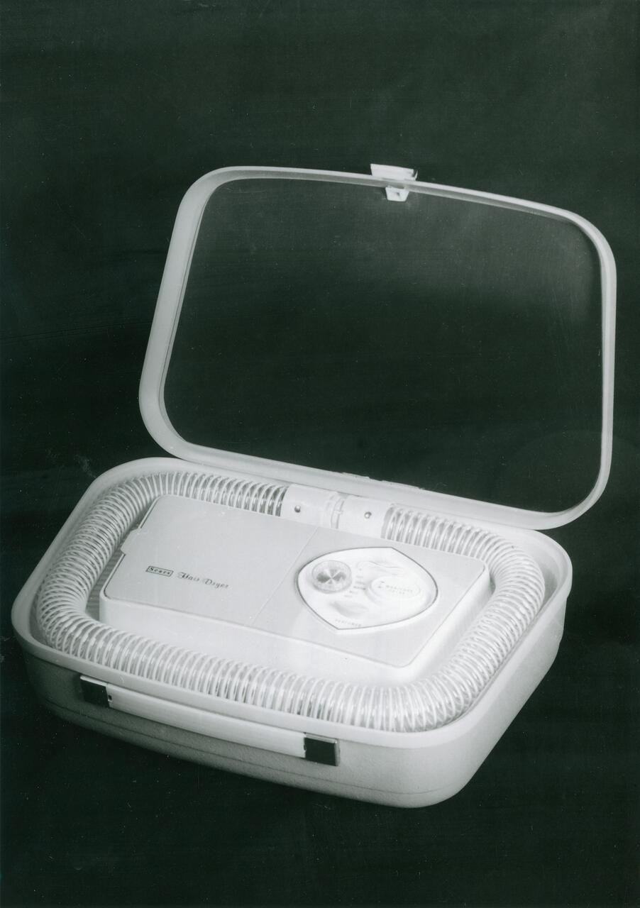 Vintage hair dryer in an open hard case with a hose and control dial, set against a dark background.