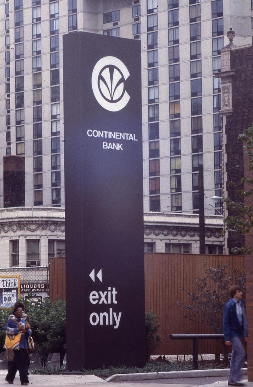 Continental Bank sign with logo and "exit only" text in front of a high-rise building. Two people walk nearby, with greenery and urban elements in the background.