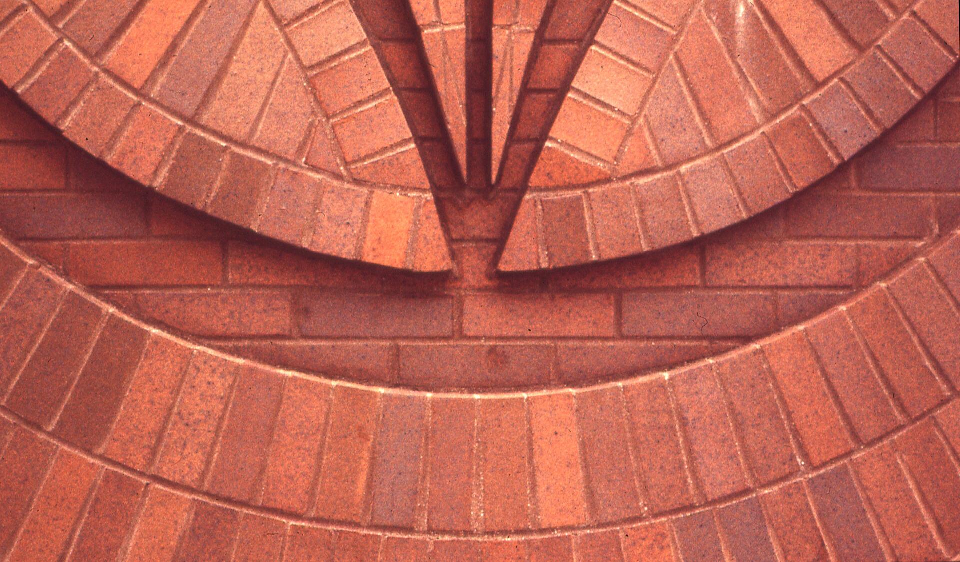 Abstract geometric pattern of interlocking red bricks forming concentric circular shapes and V-like angles.