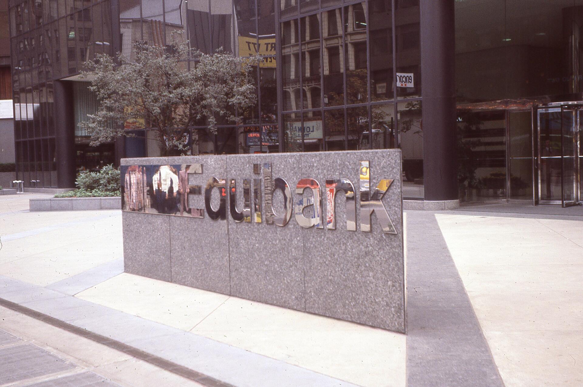 Granite wall with mirrored "Equibank" logo in an urban plaza, reflecting nearby buildings and trees.