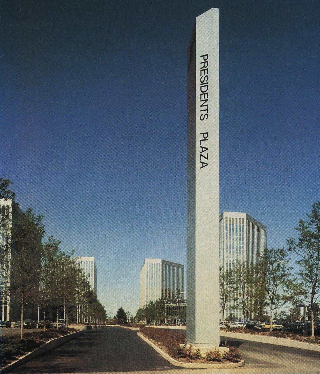 Tall stone sign with "Presidents Plaza" text stands in a landscaped area between modern glass buildings, overlooking a road flanked by trees under a clear blue sky.