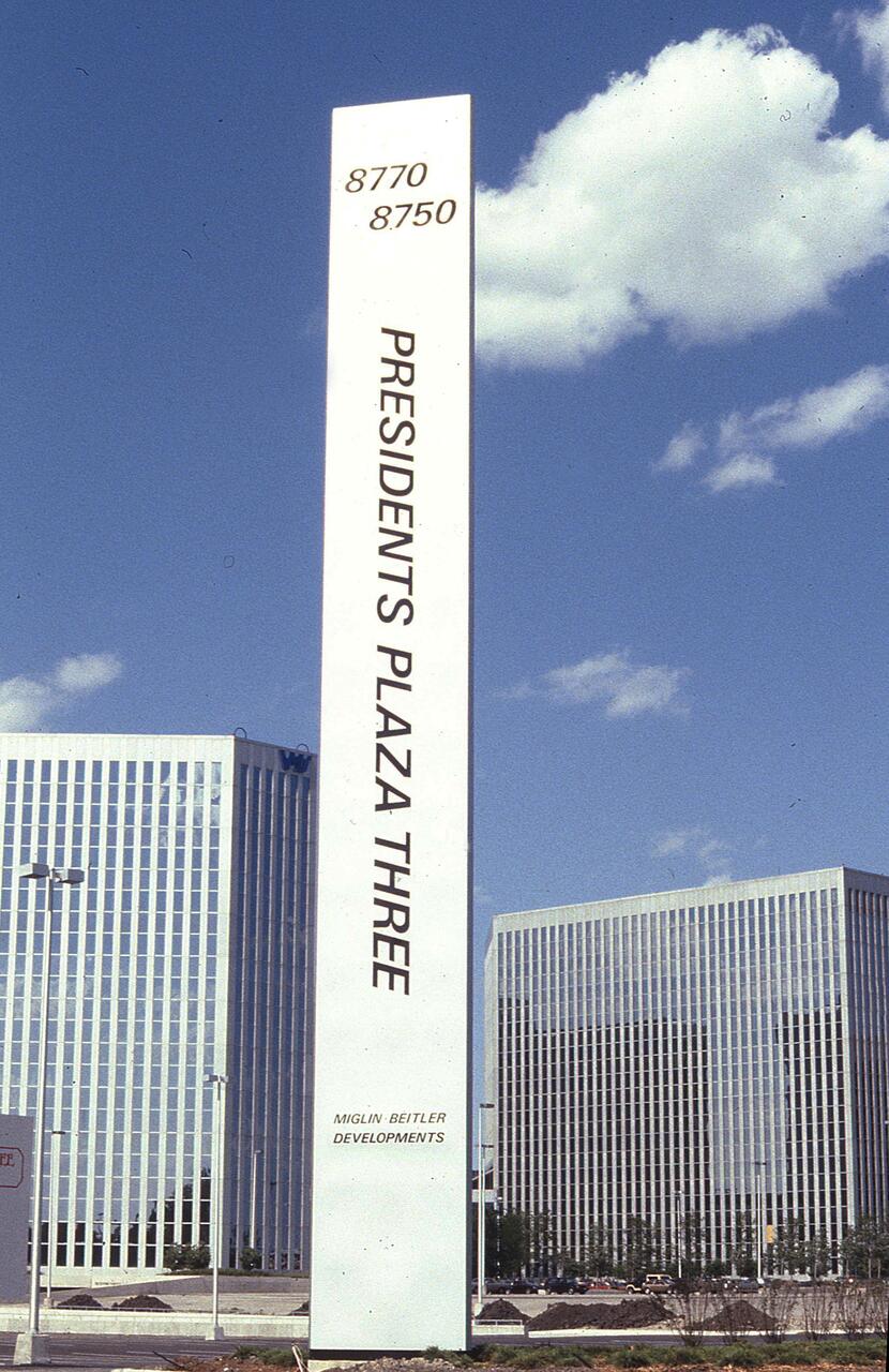 Tall vertical sign reading "Presidents Plaza Three" against a backdrop of two modern office buildings with a clear blue sky and scattered clouds.