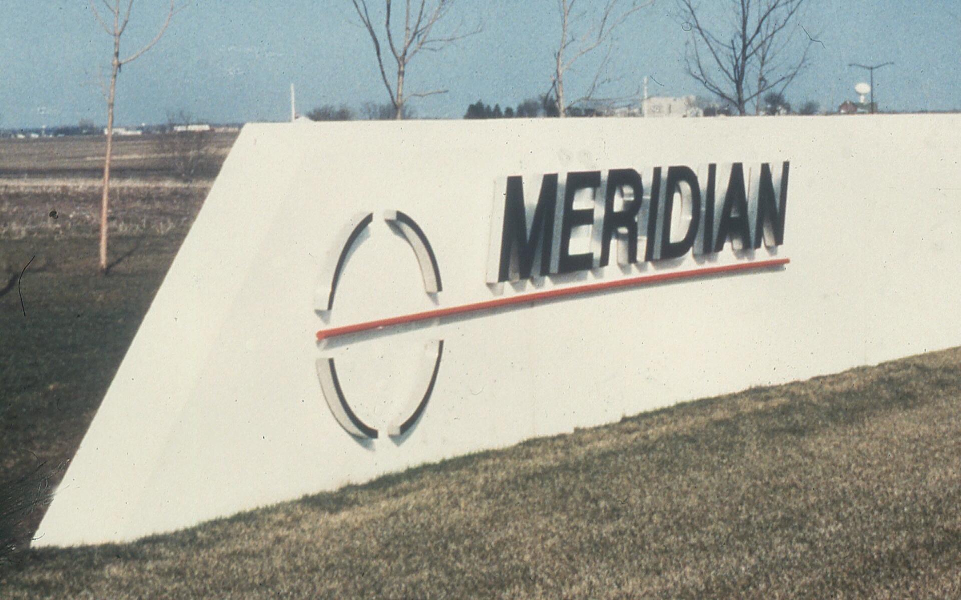 White sign displaying the word "MERIDIAN" in black letters with a red line beneath, set against a grassy field with trees and a clear sky in the background.