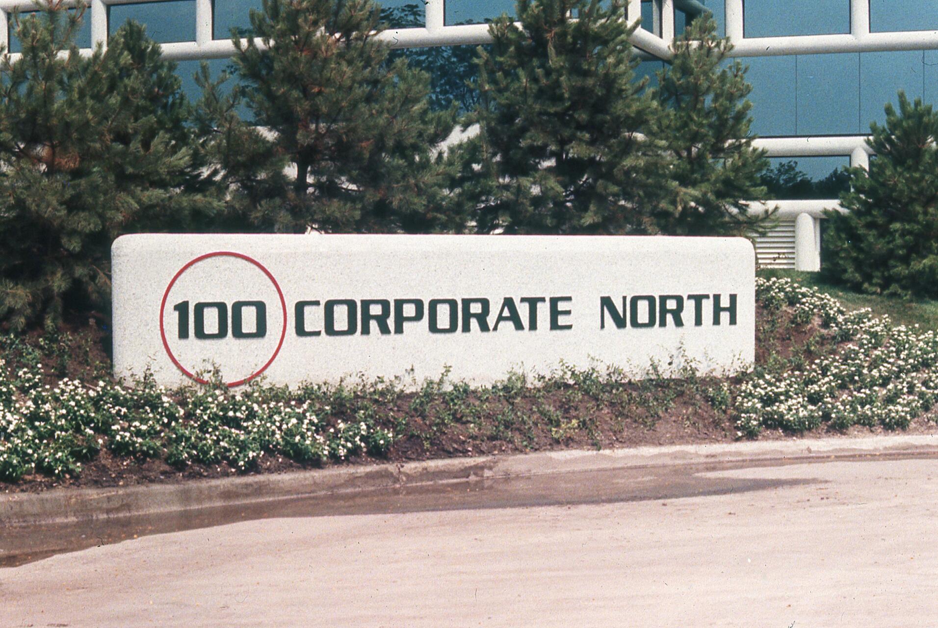 Sign reading "100 Corporate North" with a circle around "100," set in front of bushes and trees.