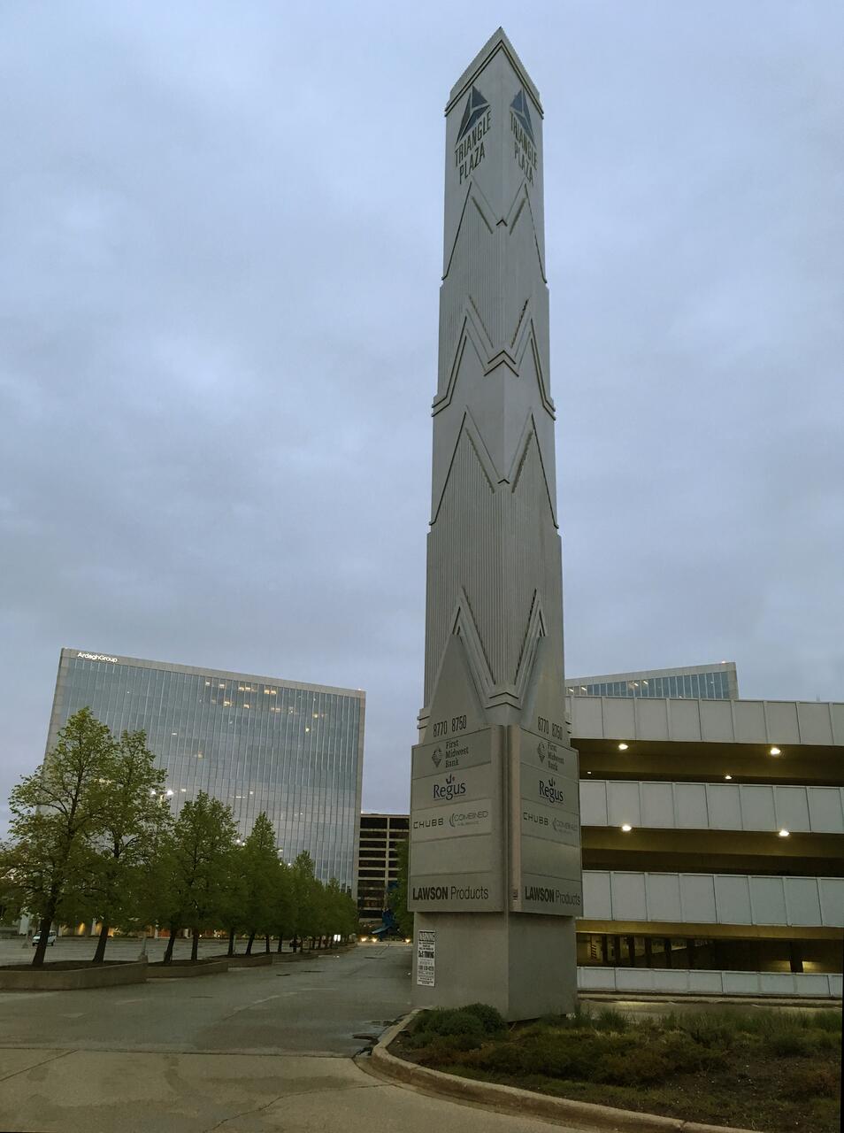 Tall, geometric monument inscribed with "Triangle Plaza" stands in a business park with modern office buildings and parking structure under an overcast sky.