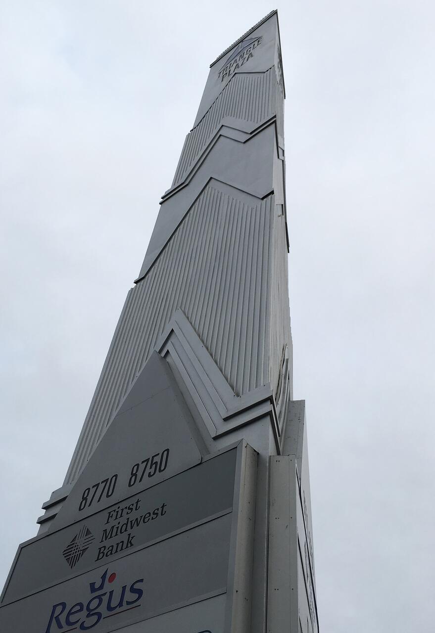 Tall, gray sign tower with geometric patterns against a cloudy sky, displaying business names including First Midwest Bank and Regus.