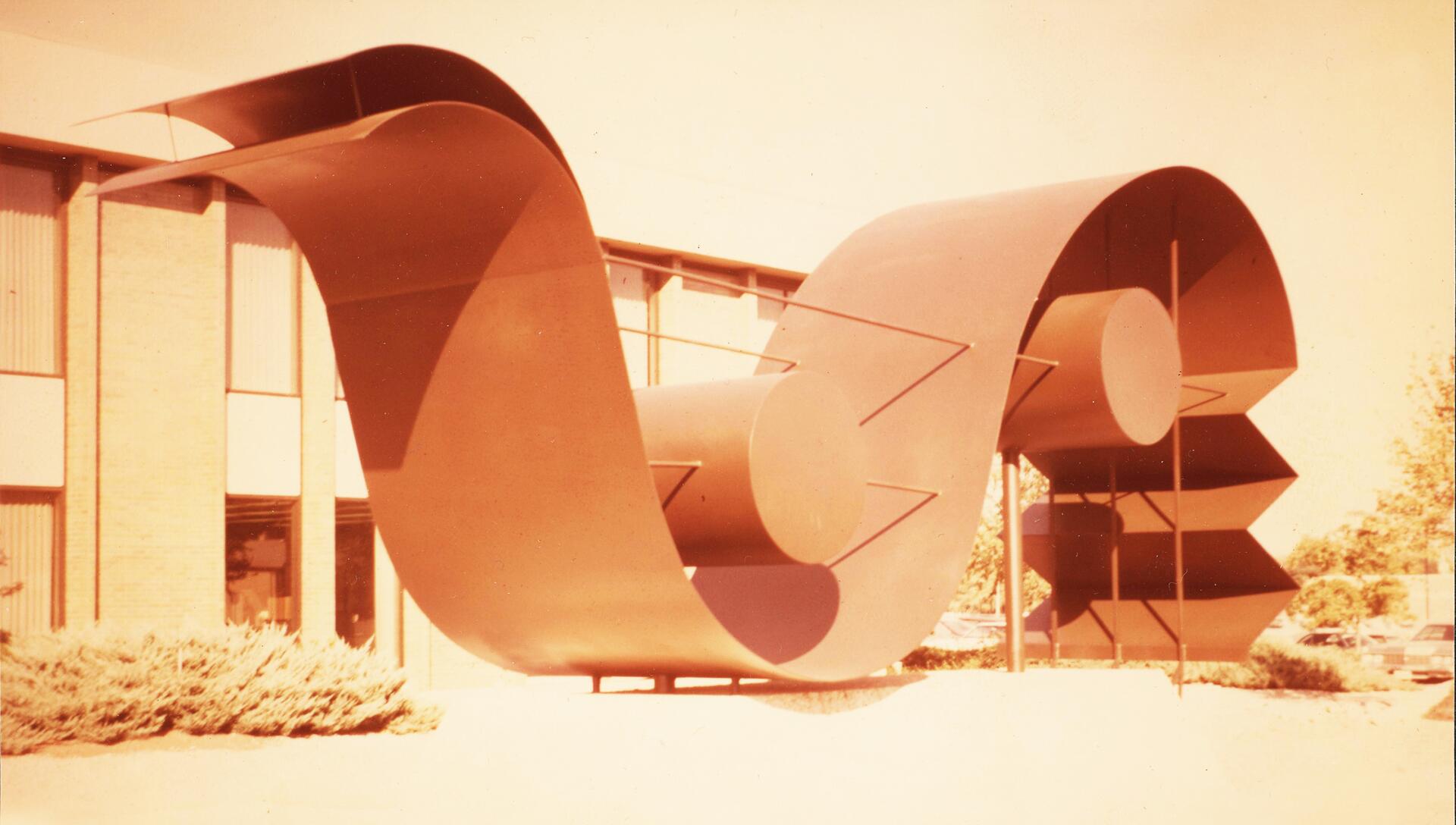 Large abstract metal sculpture with curved and circular elements, set in front of a brick building with vertical windows. Bushes are visible at the base. The image has a warm vintage tone.