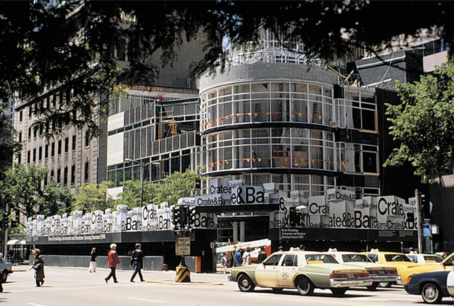 Street view of a multi-story building under construction, wrapped with Crate & Barrel signage. People walk on the sidewalk and yellow taxis are on the road. Trees partially frame the scene.