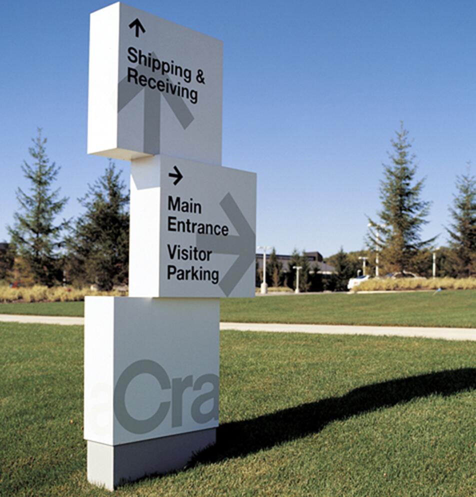 Directional signpost on a lawn with arrows pointing to "Shipping & Receiving," "Main Entrance," and "Visitor Parking." Trees and a clear sky are in the background.