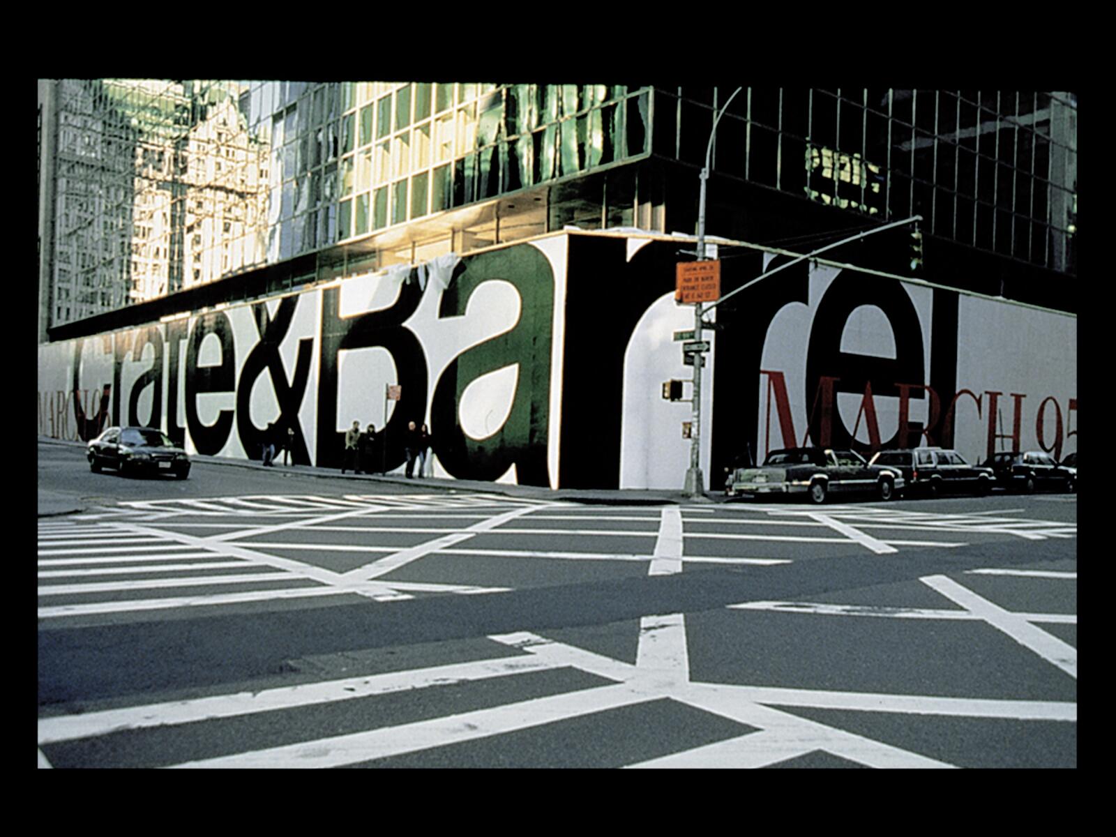 Large street corner billboard with bold letters "Crate & Barrel" on a busy intersection. Cars and pedestrians are visible, with tall buildings in the background.
