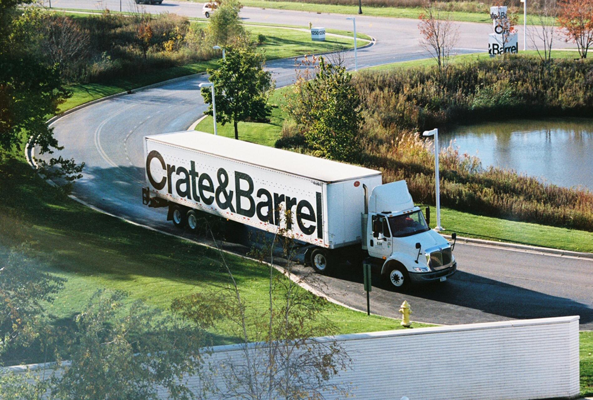 Truck with "Crate & Barrel" logo on the side driving on a curved road, surrounded by grass, trees, and a small pond, under clear blue skies.